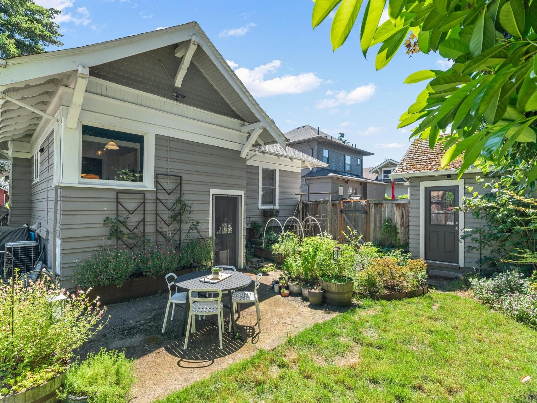 A quaint backyard features a small patio with a table and two chairs. Potted plants and greenery surround the seating area. A gray house with white trim is in the background, and a wooden shed is visible to the right. The sky is clear and sunny.