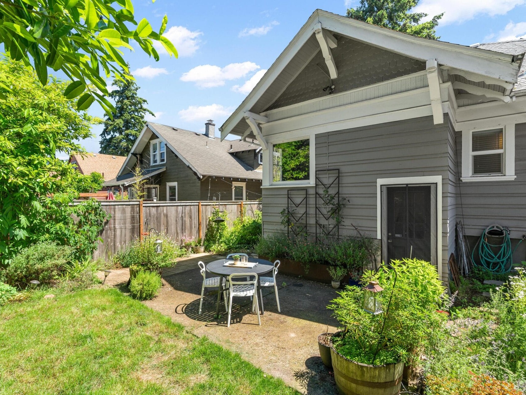 A quaint backyard with a gray house featuring white trim. Theres a small patio with a round table and two chairs, surrounded by lush greenery and plants. A garden hose is coiled by the wall. The sky is clear and sunny.