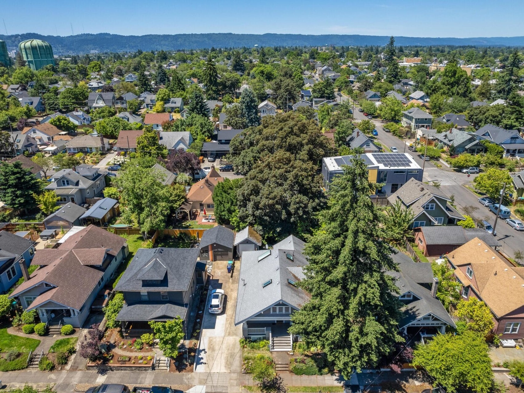 Aerial view of a suburban neighborhood with tree-lined streets and numerous houses. Some rooftops have solar panels. The area is dense with greenery, and a few taller structures are visible in the distance under a clear blue sky.