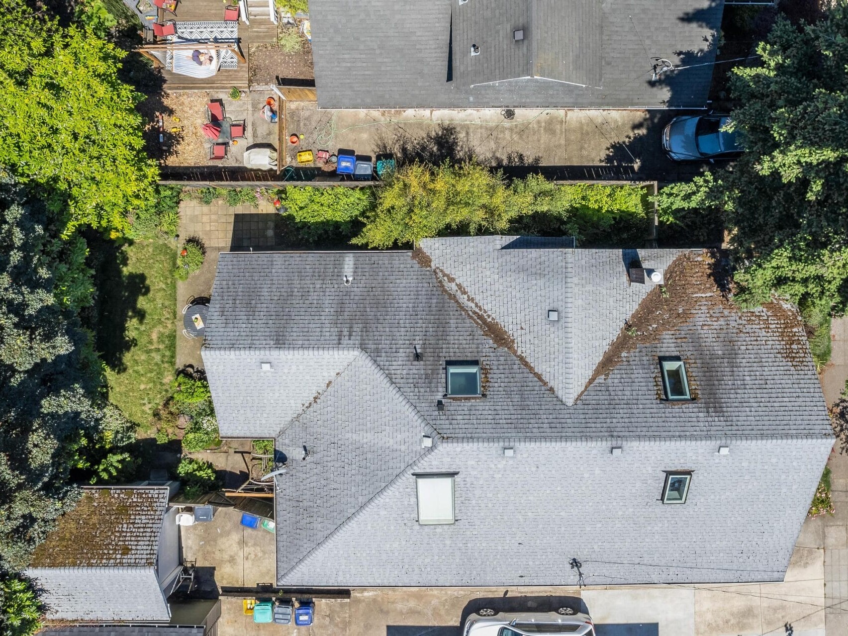 Aerial view of two houses with gray roofs, surrounded by trees and yards. One house has skylights and a driveway with parked cars. Various items like bins and equipment are visible in the backyards.