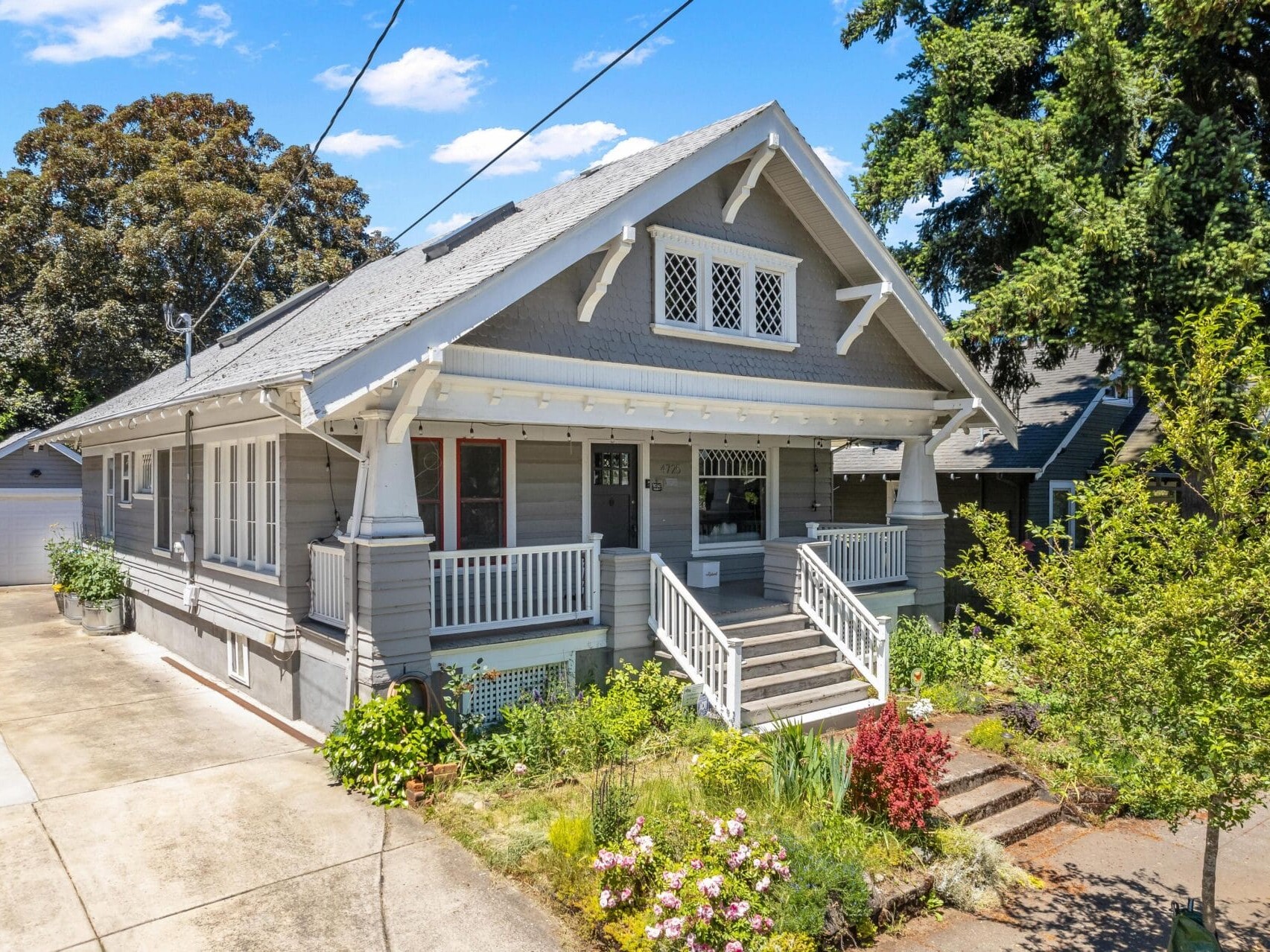 A charming gray craftsman-style house with a gabled roof, white trimmings, and a spacious front porch. It is surrounded by green shrubs, colorful flowers, and tall trees under a clear blue sky. A driveway leads to a garage in the back.