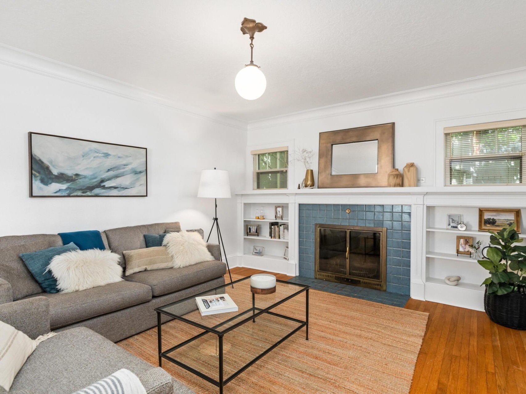 A cozy living room with a gray couch adorned with pillows, a coffee table, and a blue-tiled fireplace. Shelves with decor items flank the fireplace. A large painting and a floor lamp add to the decor, with hardwood floors and a warm atmosphere.
