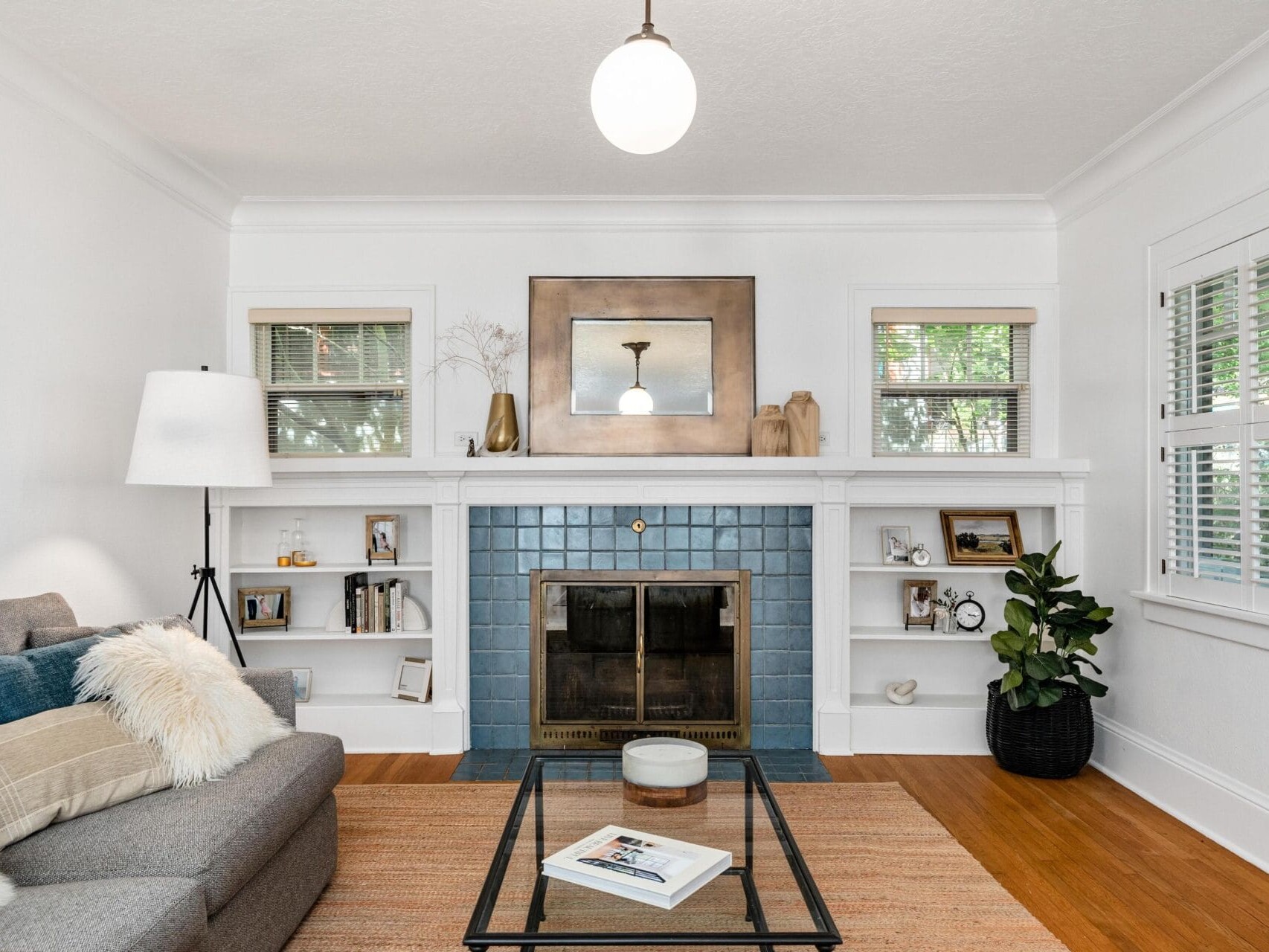 A cozy living room with a gray sofa, fluffy pillows, and a glass coffee table. A fireplace with blue tiles is set in a white mantel, flanked by two windows. Decor items and a potted plant add charm to the space. A light fixture hangs above.