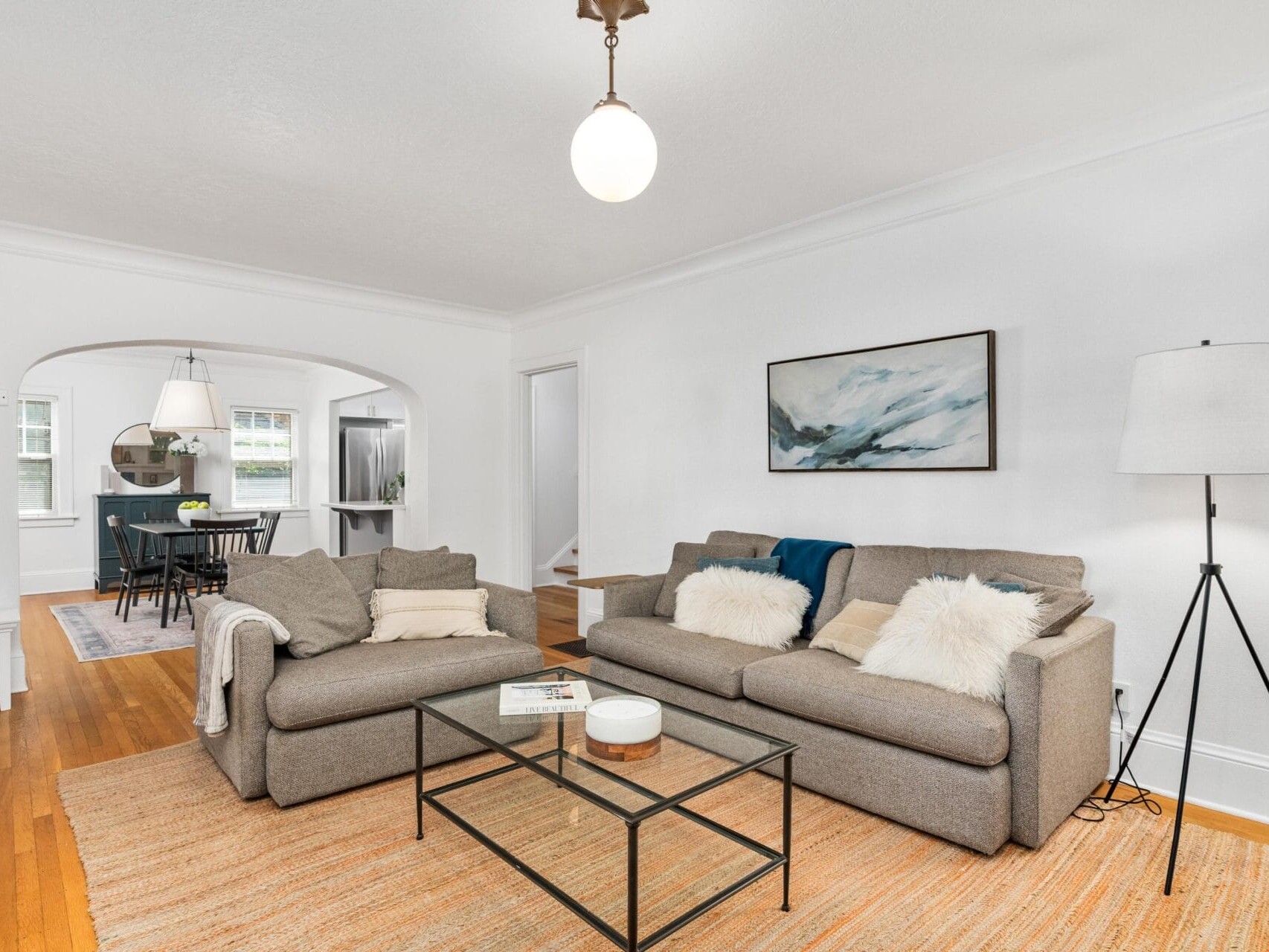 A cozy living room with two beige sofas, a glass coffee table, and a standing lamp on a beige rug. A landscape painting hangs on the wall. The room opens into a dining area with a table and chairs visible through an archway.