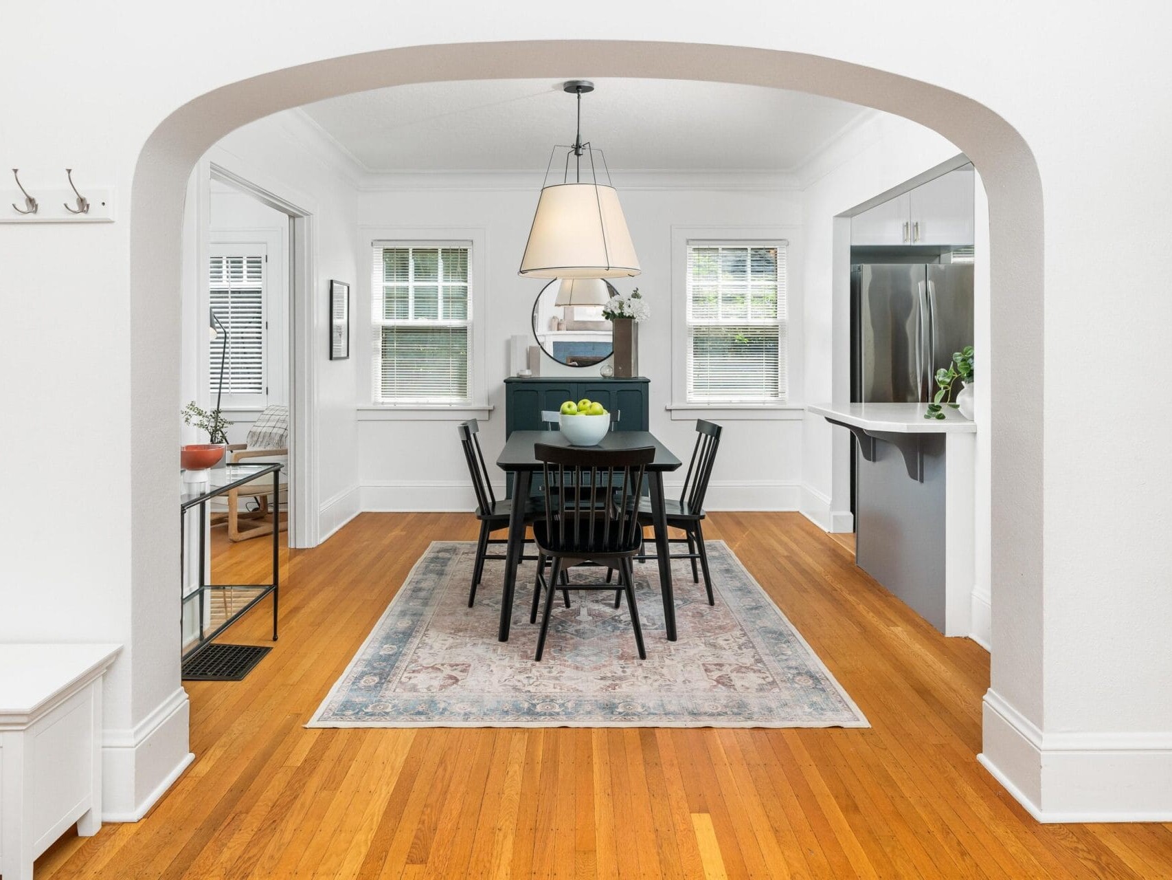 A dining room with a wooden floor and a blue rug features a black table and chairs. A pendant light hangs above. The room has white walls, windows, plants, and arched entrances. Kitchen appliances are partially visible on the right.