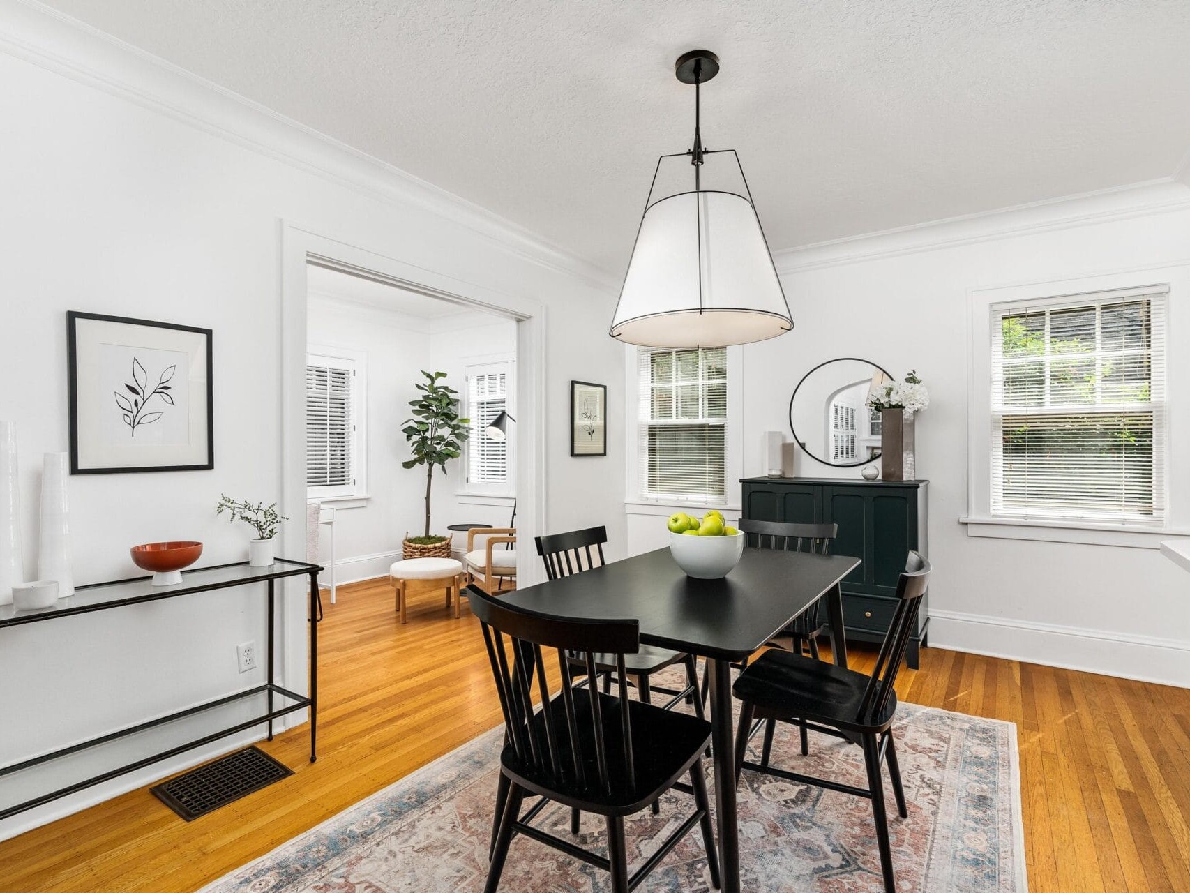 A dining room with a black table and four chairs on a patterned rug. A large pendant light hangs above the table, which features a bowl of green apples. The room has hardwood floors, a window, and minimal decor, including a small console table and wall art.