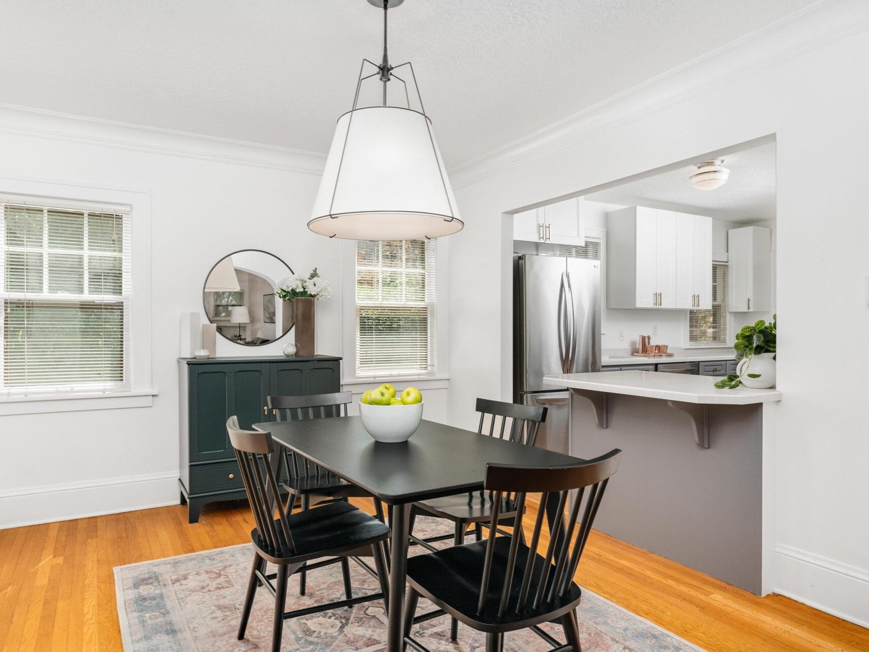 A bright dining area with a black table and four chairs on a patterned rug. A bowl of green apples is on the table. A large pendant light hangs above. A black cabinet with a round mirror is against the wall. The kitchen is visible in the background.