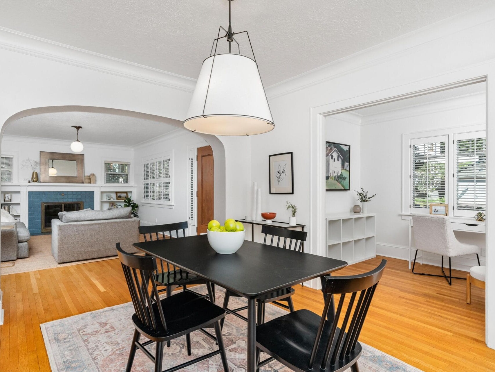 A bright dining room with a black table and chairs, a white bowl of green apples, and a large pendant light above. Adjacent rooms include a living area with a fireplace and a small study with a desk. Hardwood floors and white walls throughout.