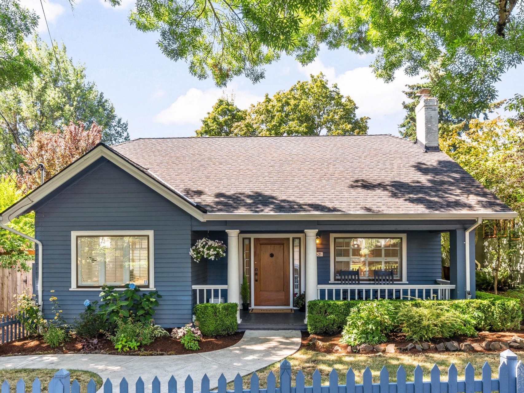 A charming blue cottage with a white picket fence, surrounded by lush greenery. The house features a small porch with hanging plants, large windows, and a chimney. The yard is neatly landscaped with shrubs and a curved walkway.