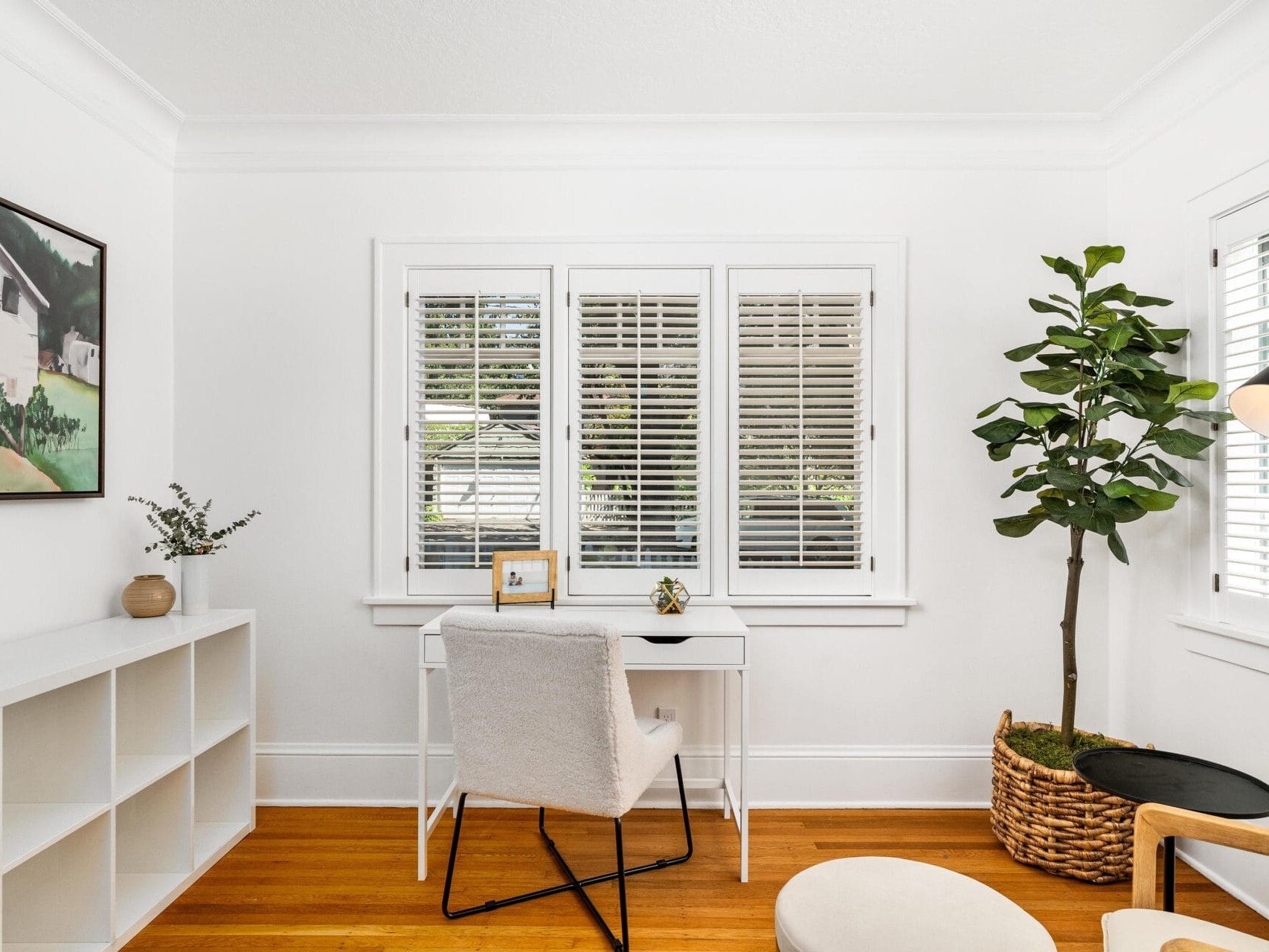 A bright home office with a white desk and chair facing a window with blinds. A large potted plant sits on the right, and a small framed picture and succulent are on the desk. A white shelving unit is on the left wall.