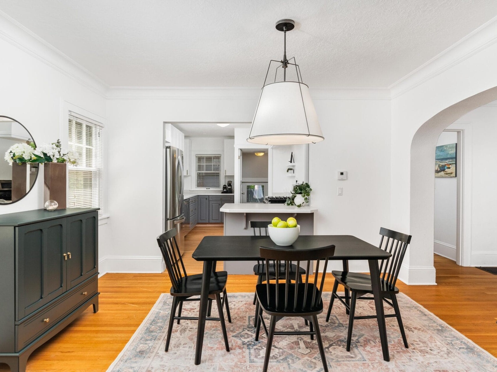 Dining room with a black table and four chairs on a patterned rug, under a large white lamp. A bowl of green apples sits on the table. In the background, theres a view into a kitchen with blue cabinets. A dark cabinet with a round mirror is on the left.