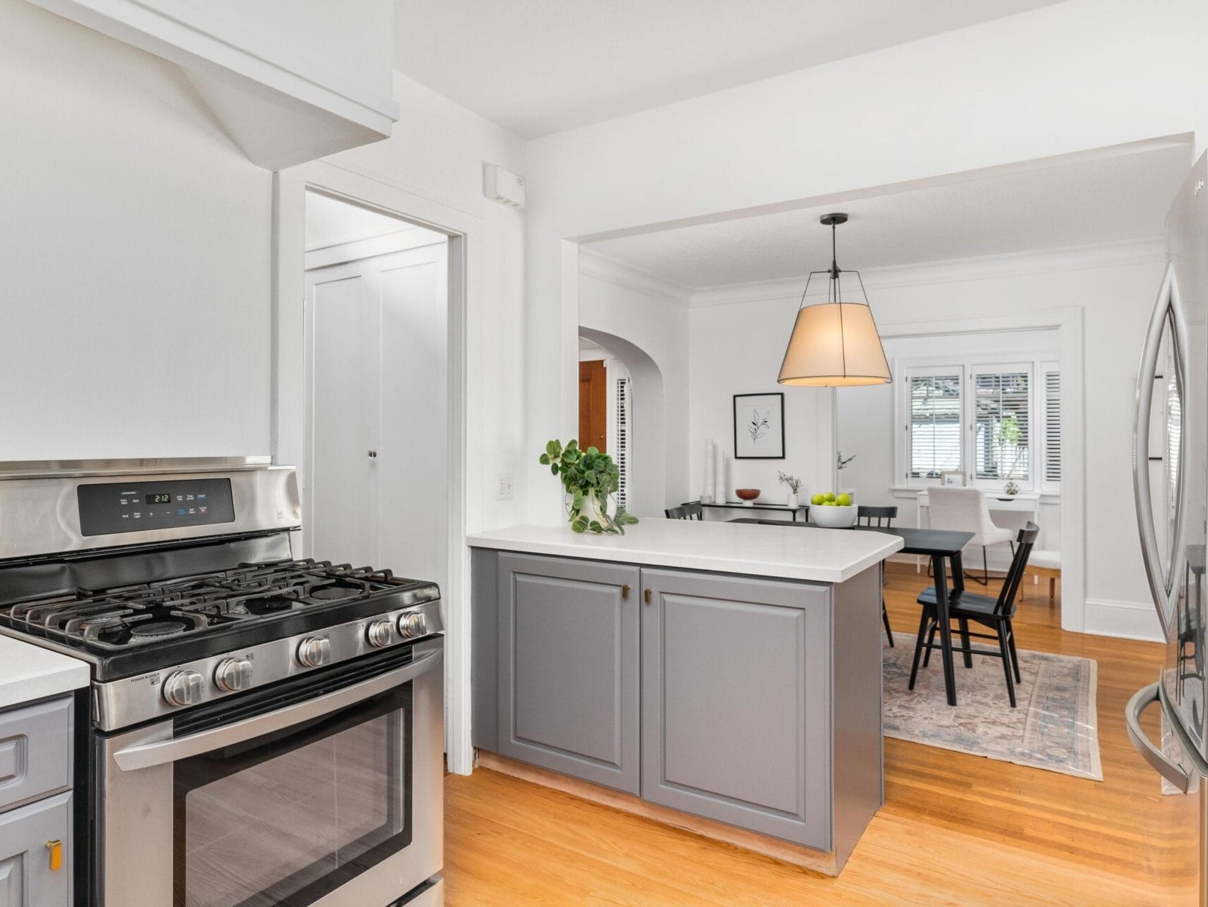 A modern kitchen and dining area with stainless steel appliances, grey cabinetry, and light wood flooring. A dining table with chairs and a large pendant lamp is visible in the adjacent room. White walls and windows enhance the bright space.