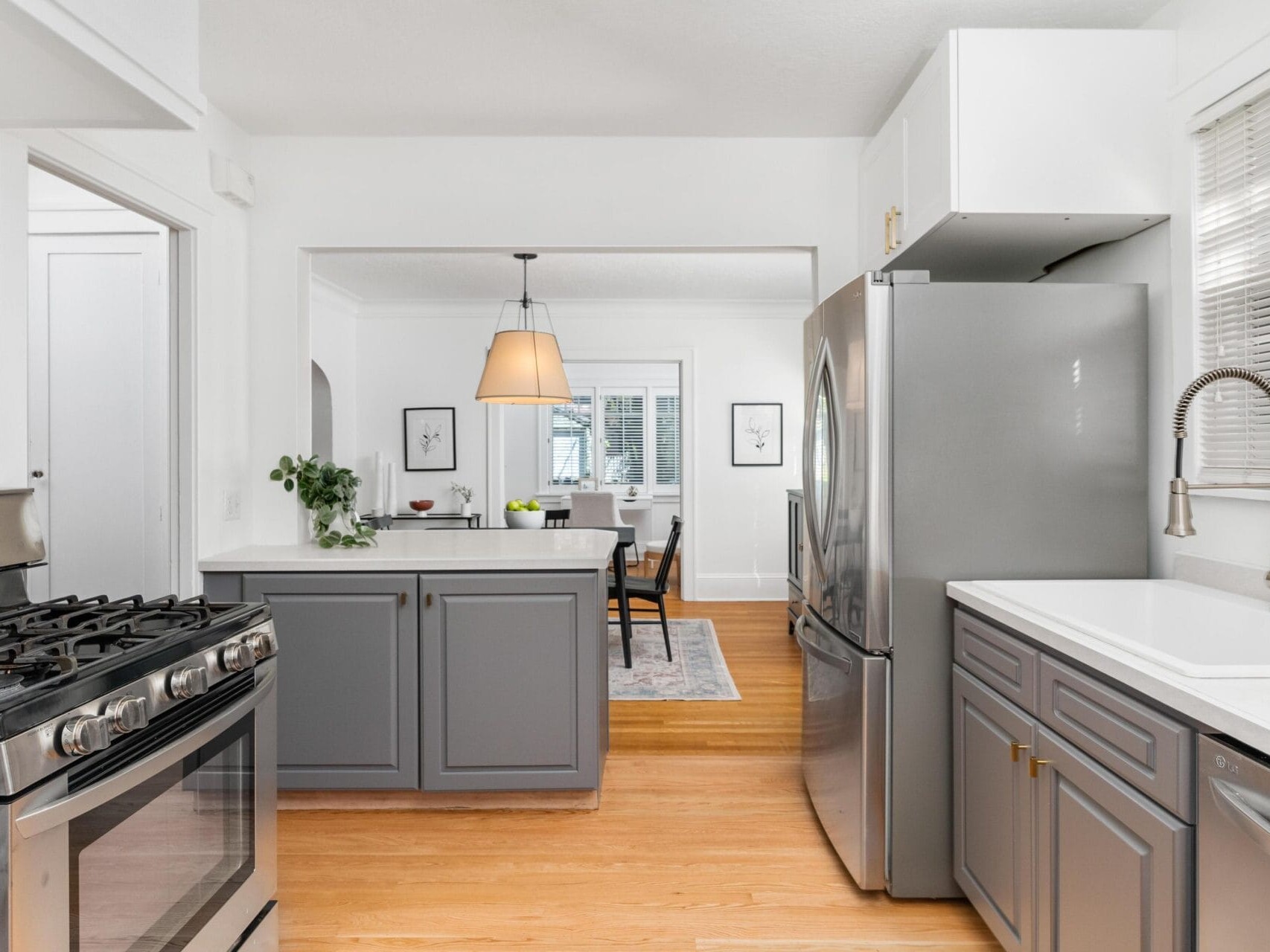 A modern kitchen with stainless steel appliances, including a stove and refrigerator. Gray cabinets complement white countertops. A view into the dining area shows a table with chairs, a pendant light, and wall art. Light wooden floors unify the spaces.