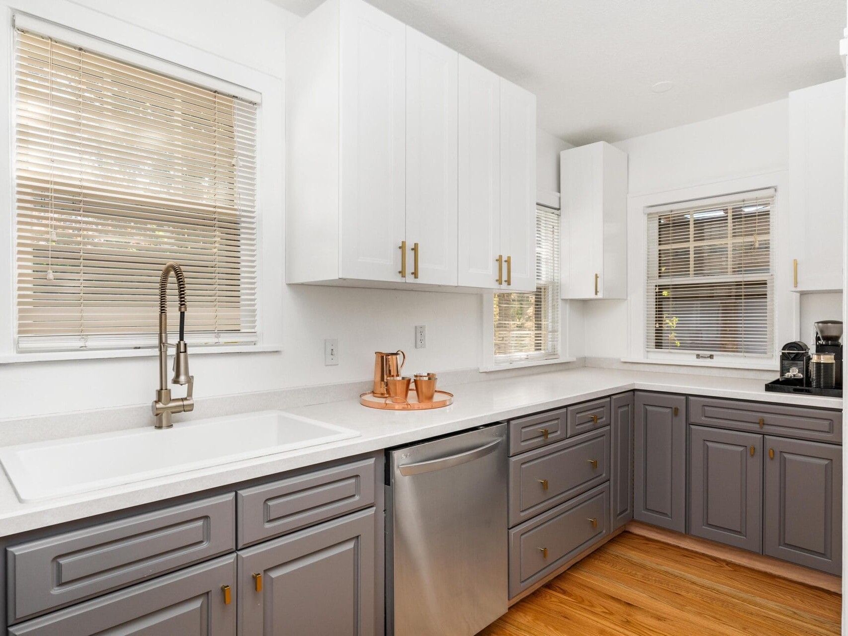 A modern kitchen with gray cabinetry, white countertops, and stainless steel appliances. The space features a large sink, copper utensils, hardwood flooring, and windows with blinds, allowing natural light to illuminate the room.