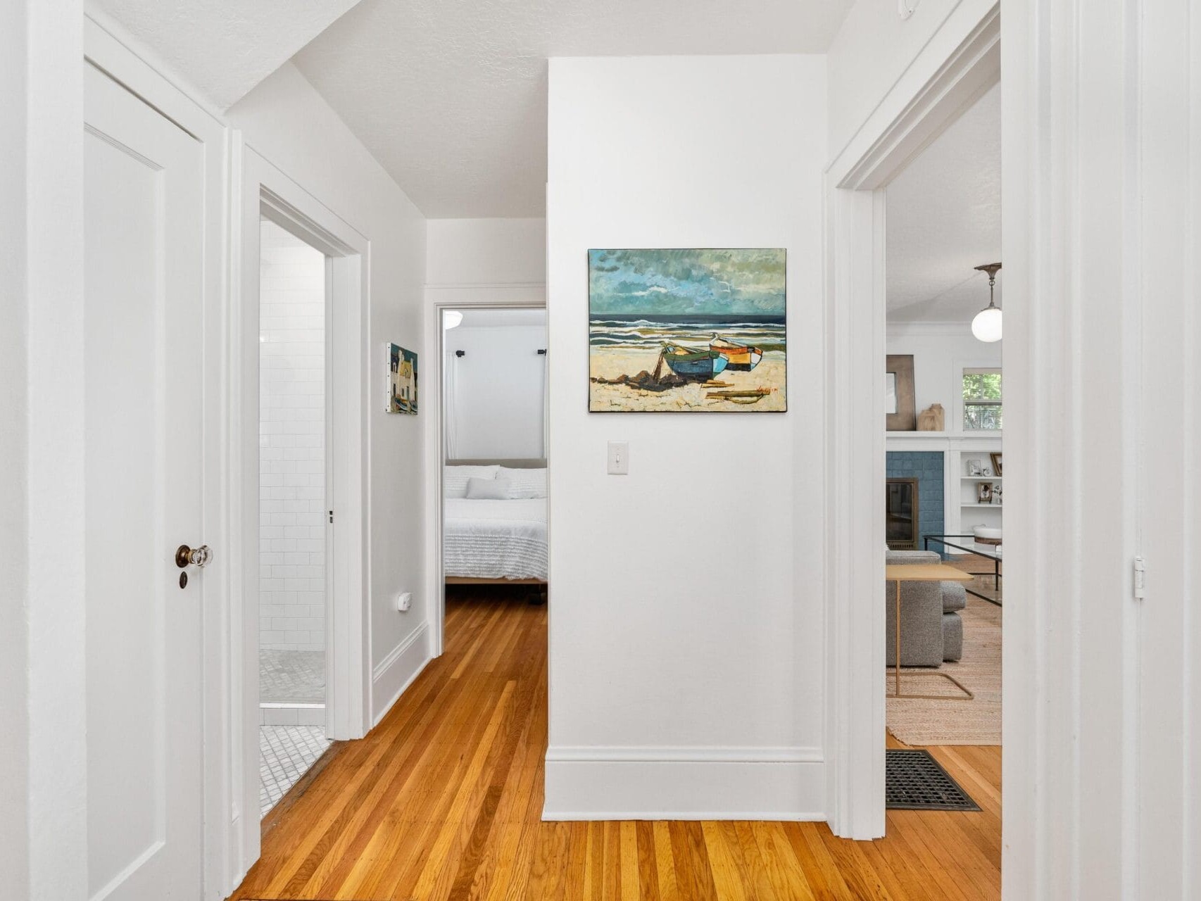 A hallway with wooden floors leads to three rooms. On the left, theres a white-tiled bathroom. Straight ahead, theres a bedroom with a white bedspread. On the right, a living room is visible. A beach painting hangs on the wall.
