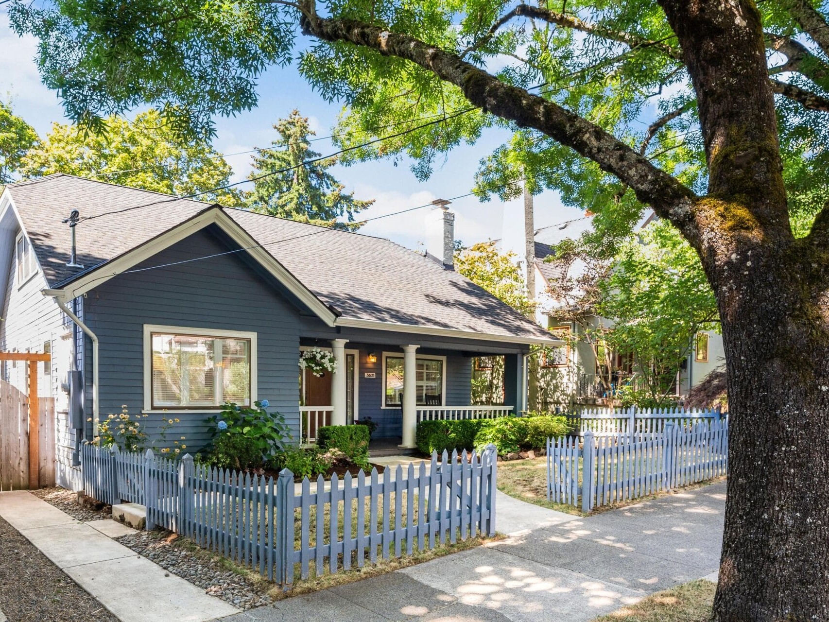 A charming blue house with white trim sits behind a picket fence. The front yard features green bushes, and a large tree provides shade. The house has a welcoming porch with white railings and is set in a neighborhood with greenery and trees.