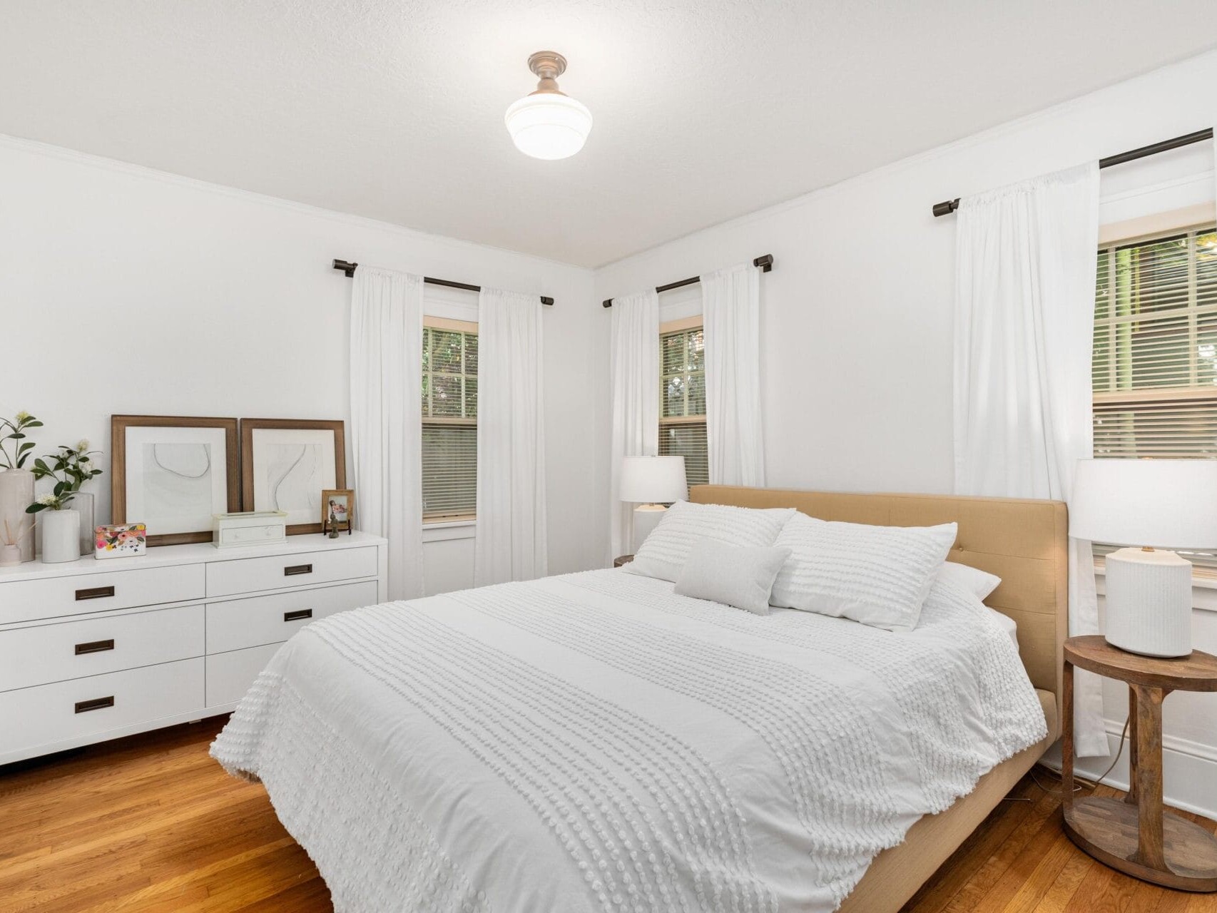 A bright bedroom with a large bed covered in white bedding, two white pillows, and a beige headboard. A wooden side table with a lamp is beside the bed. A white dresser with decor is against the wall. Two windows with white curtains let in natural light.