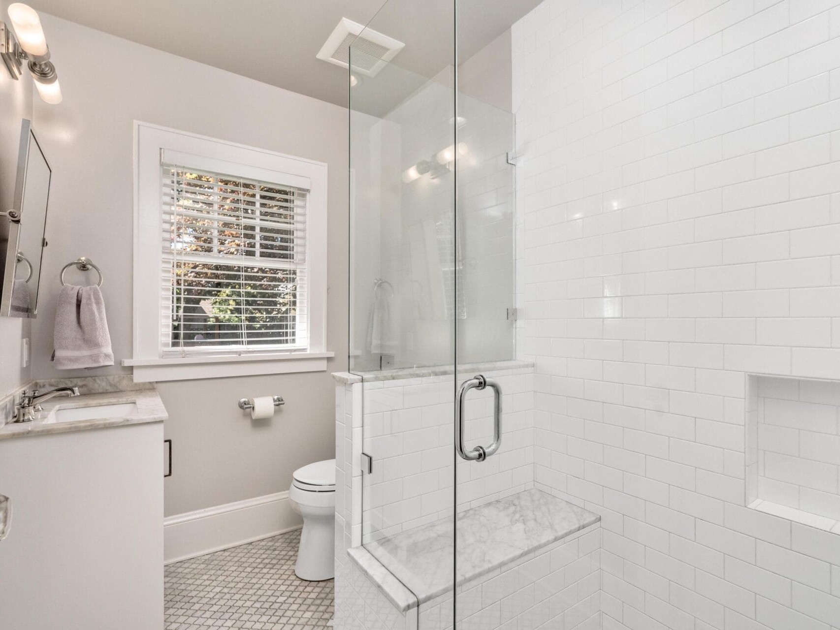 A modern bathroom with a glass-enclosed shower, white subway tiles, a small window with blinds, a towel hanging by the sink, and a toilet. The floor features a light gray geometric tile pattern.