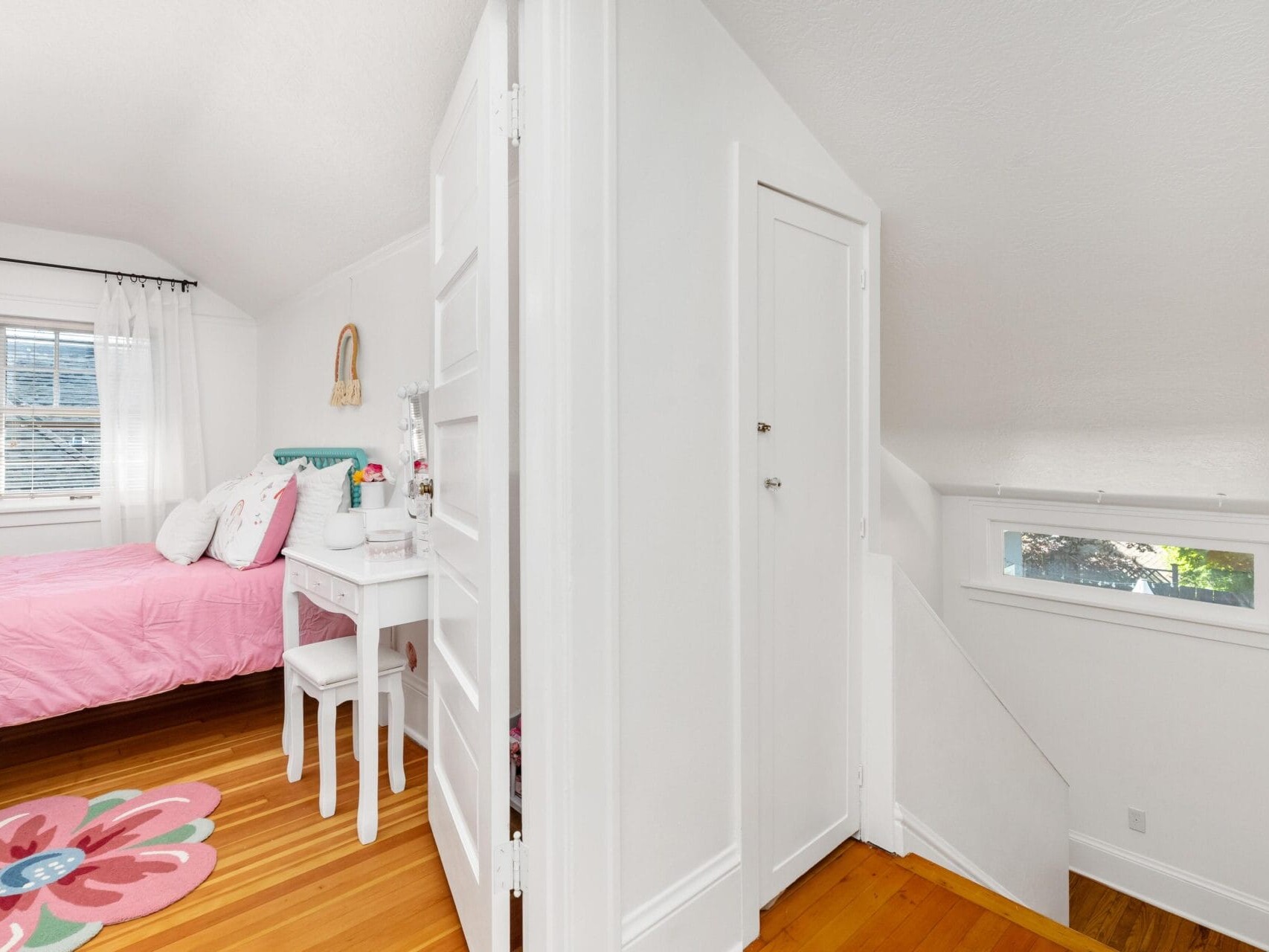 A bright bedroom with a pink bedspread, floral rug, and white furniture, separated by a wall from a small hallway with stairs. The space is illuminated by natural light from a window.