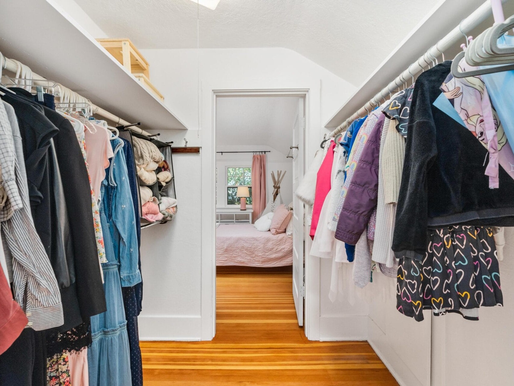 A walk-in closet with clothes hanging on both sides and a wooden floor. The closet leads to a bright bedroom with a bed covered in white and pink linens. A window is visible in the background.