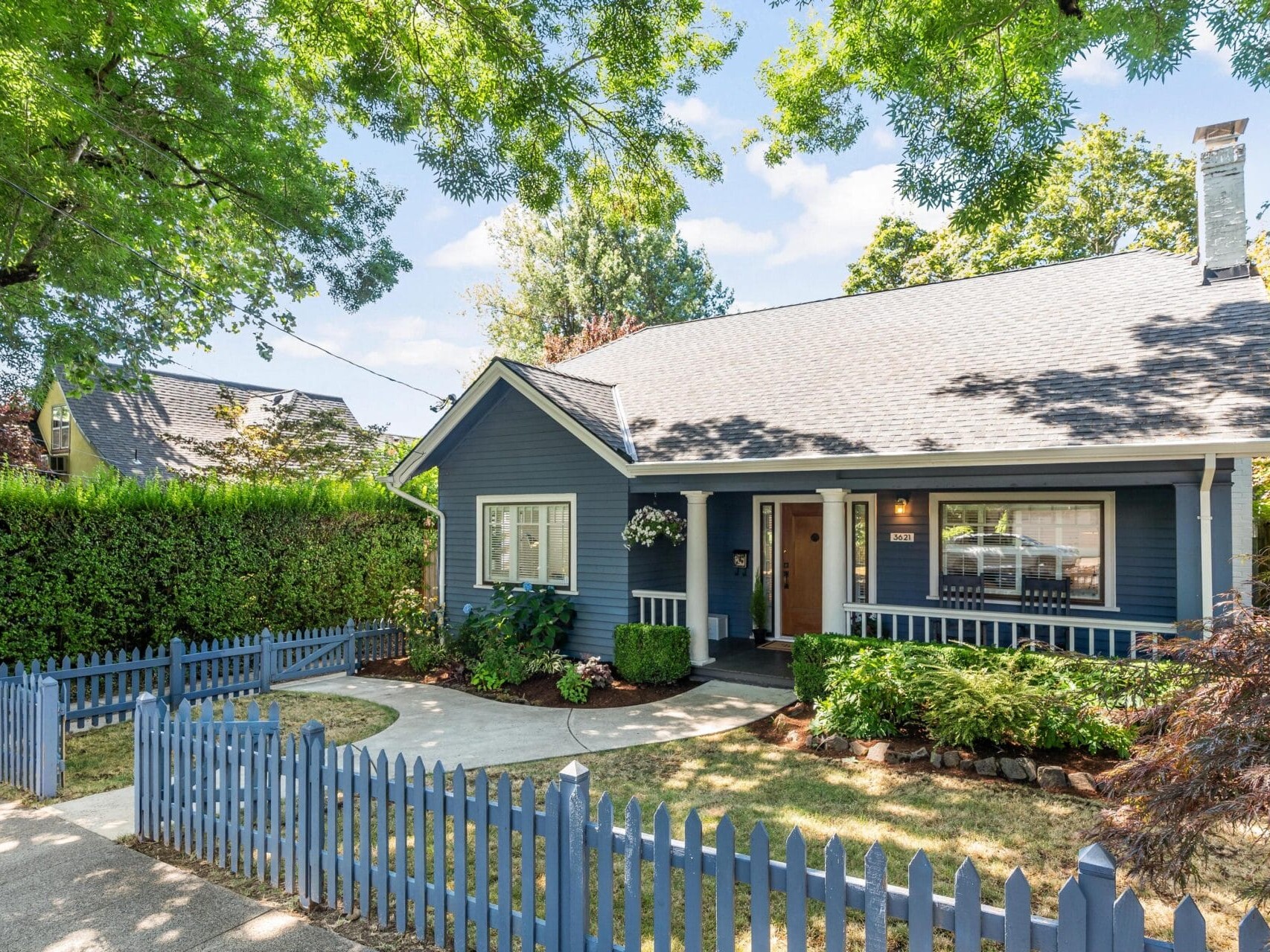 A charming blue house with a white trim stands behind a blue picket fence. It features a neatly kept lawn, a path leading to the front door, and lush green trees providing shade. The sky above is clear and sunny.