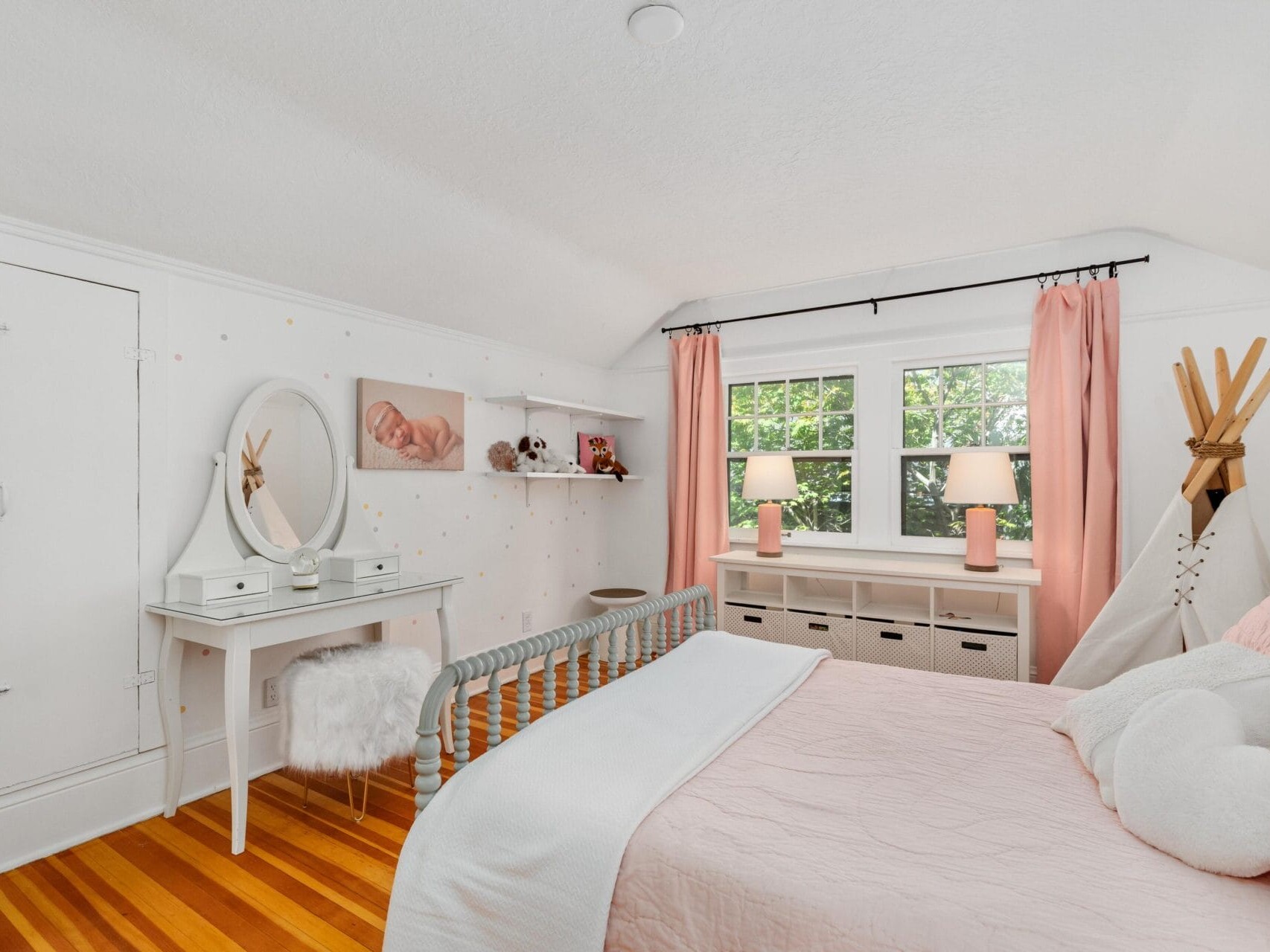 A cozy bedroom with a white and pink color scheme. It features a bed with plush pillows, a vanity with a round mirror and fur-covered stool, a teepee, shelves with decor, and large windows with pink curtains letting natural light in.