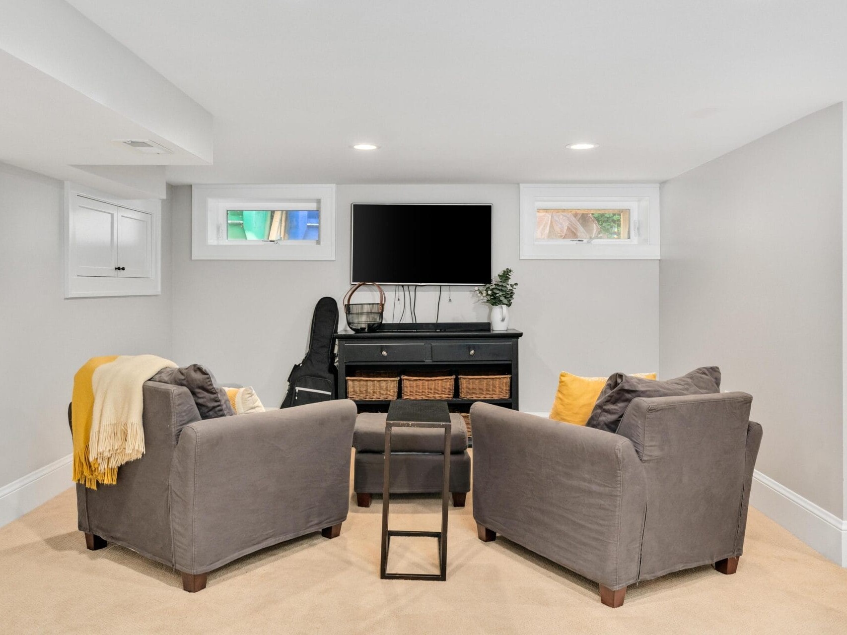 A cozy living room with two gray armchairs facing a wall-mounted TV. A small black side table is between the chairs. A cabinet with baskets and decor sits beneath the TV. Soft lighting and a beige carpet create a warm ambiance.