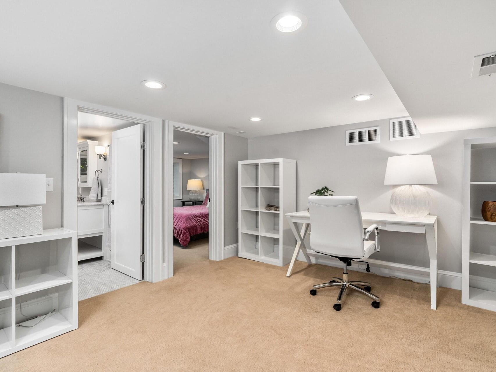 A bright, modern home office with beige carpet, featuring a white desk and office chair. Shelves line the walls, with a door leading to a bathroom on the left and a bedroom straight ahead. Ceiling lights provide illumination.