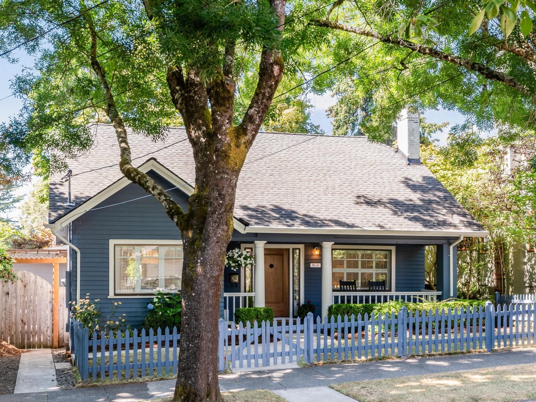 A charming blue house with a front porch, surrounded by a white picket fence and lush trees. The house has a sloped roof and large windows, casting dappled sunlight onto the front yard. A wooden gate is visible to the left.