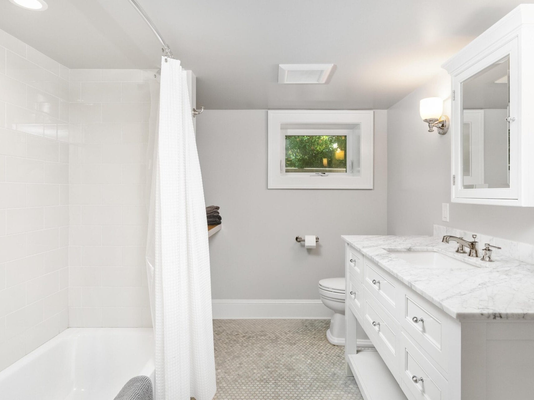 A bright, white bathroom with a bathtub and shower combo on the left, a white curtain, a vanity with a marble countertop and mirror on the right, a small window above the toilet in the center, and patterned floor tiles.
