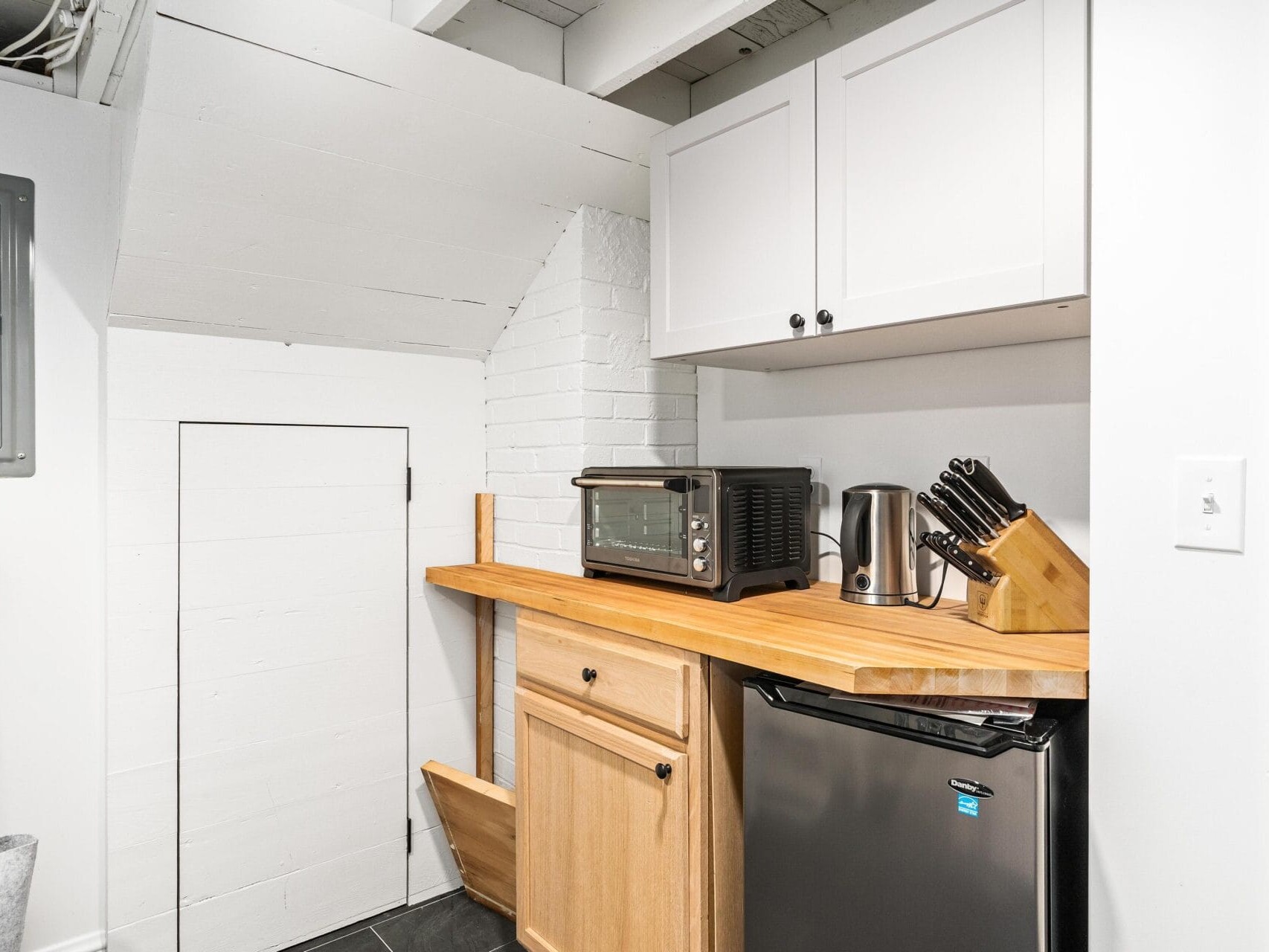 A compact kitchen corner featuring wooden countertops with a toaster oven, electric kettle, and knife block. A mini fridge is tucked beneath the counter. White cabinets are mounted above, and the space is partially under a sloped ceiling.