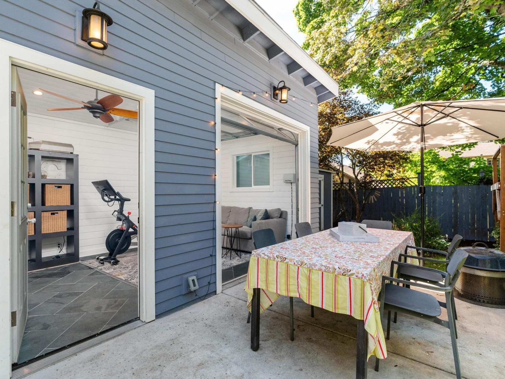 A patio with a table covered in a striped tablecloth under an umbrella, surrounded by chairs. A door opens to a room with exercise equipment, including a stationary bike. The house exterior is painted blue with trees providing shade.