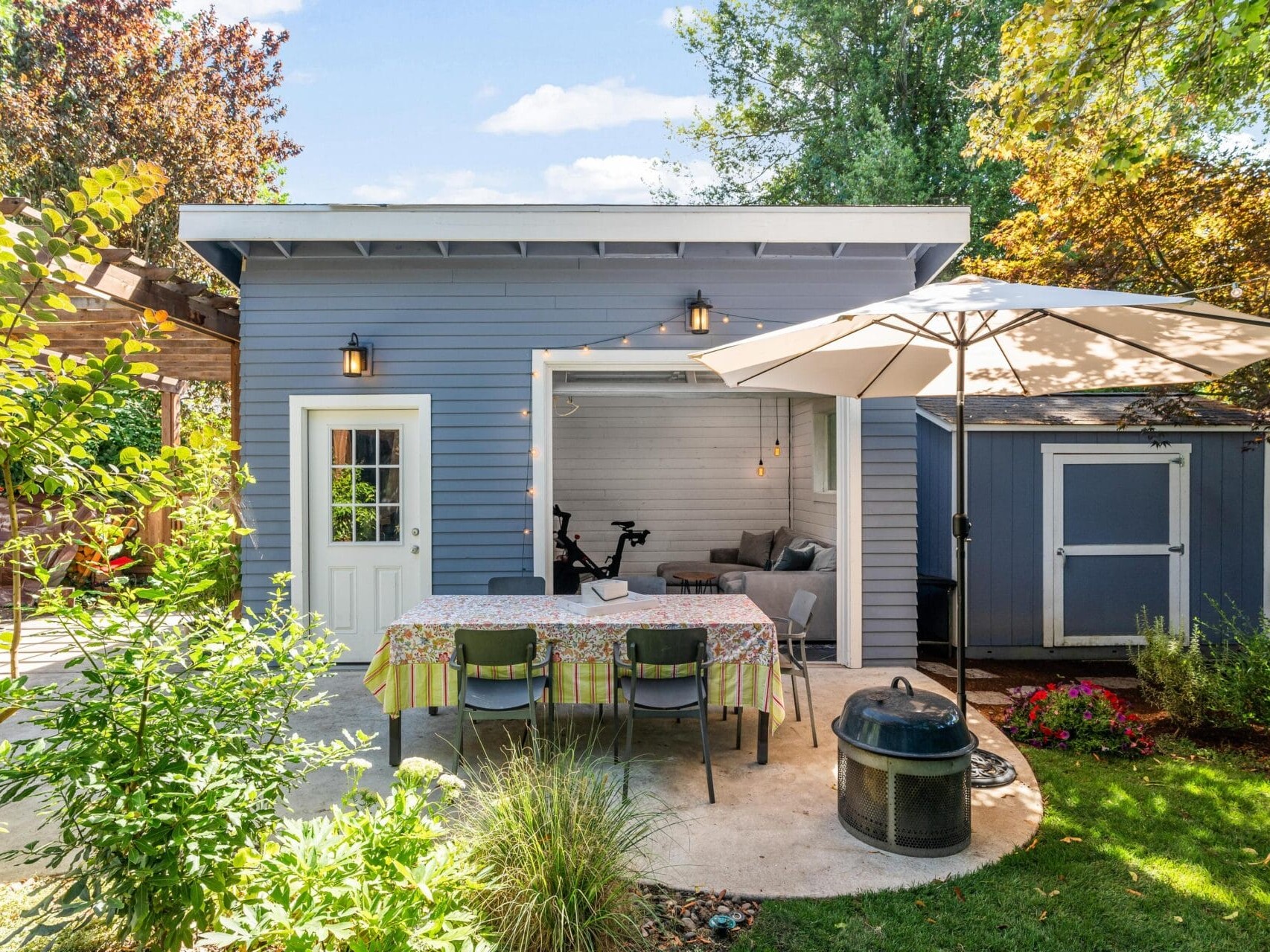 A cozy backyard patio with a small blue house, a table covered with a floral tablecloth, and chairs. An umbrella provides shade. A small shed is nearby, surrounded by lush greenery and colorful flowers. A stationary bike is visible inside.