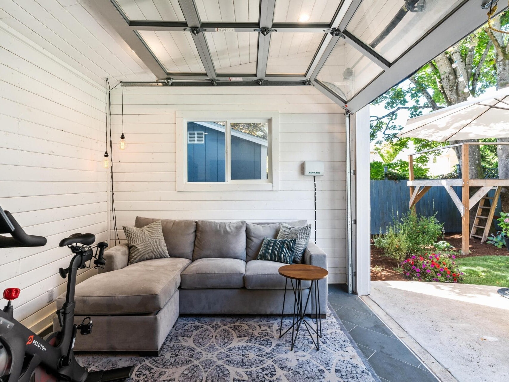 A cozy room with white wooden walls and an open garage door. Inside, theres a gray sectional sofa with cushions, a small table, and an exercise bike. Outside, a patio with plants and a large tree is visible under a clear sky.