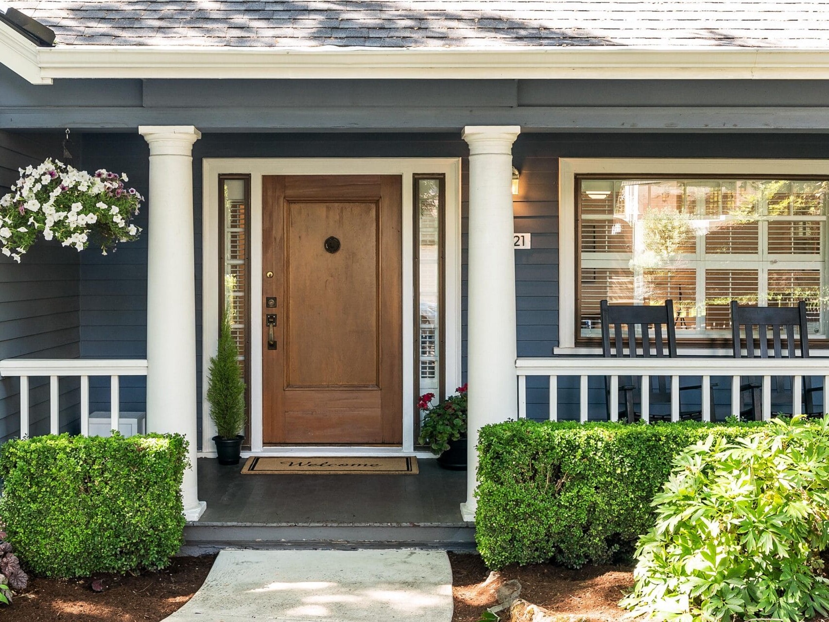 A charming house with a wooden front door flanked by two white columns. A welcome mat lies at the entrance. Theres a window with shutters on the right, a small porch with two chairs, and a hanging flower basket on the left. Green shrubs line the walkway.