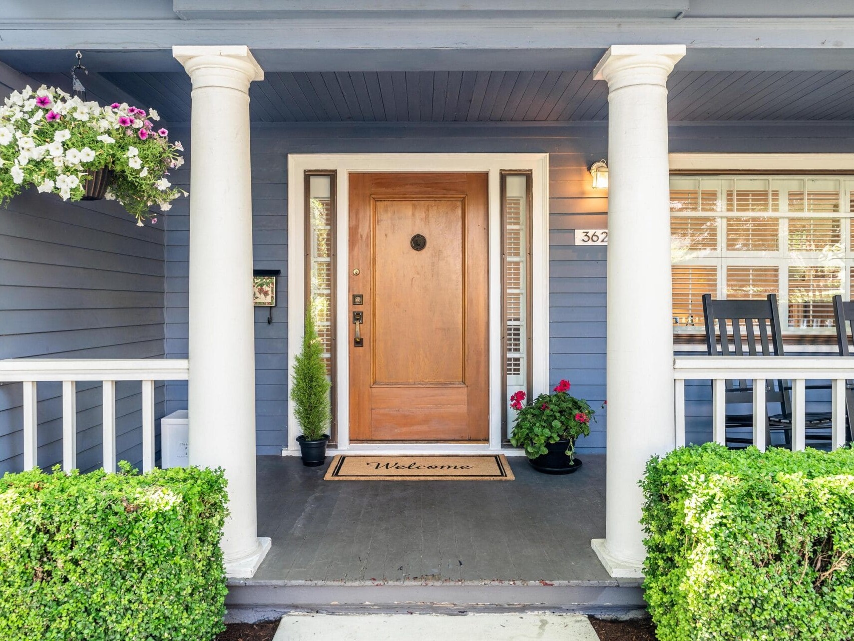 A charming front porch with a wooden door, white pillars, and a Welcome doormat. There are potted plants on either side of the door and a hanging basket with flowers. A row of black rocking chairs sits on the porch. The house is painted blue.