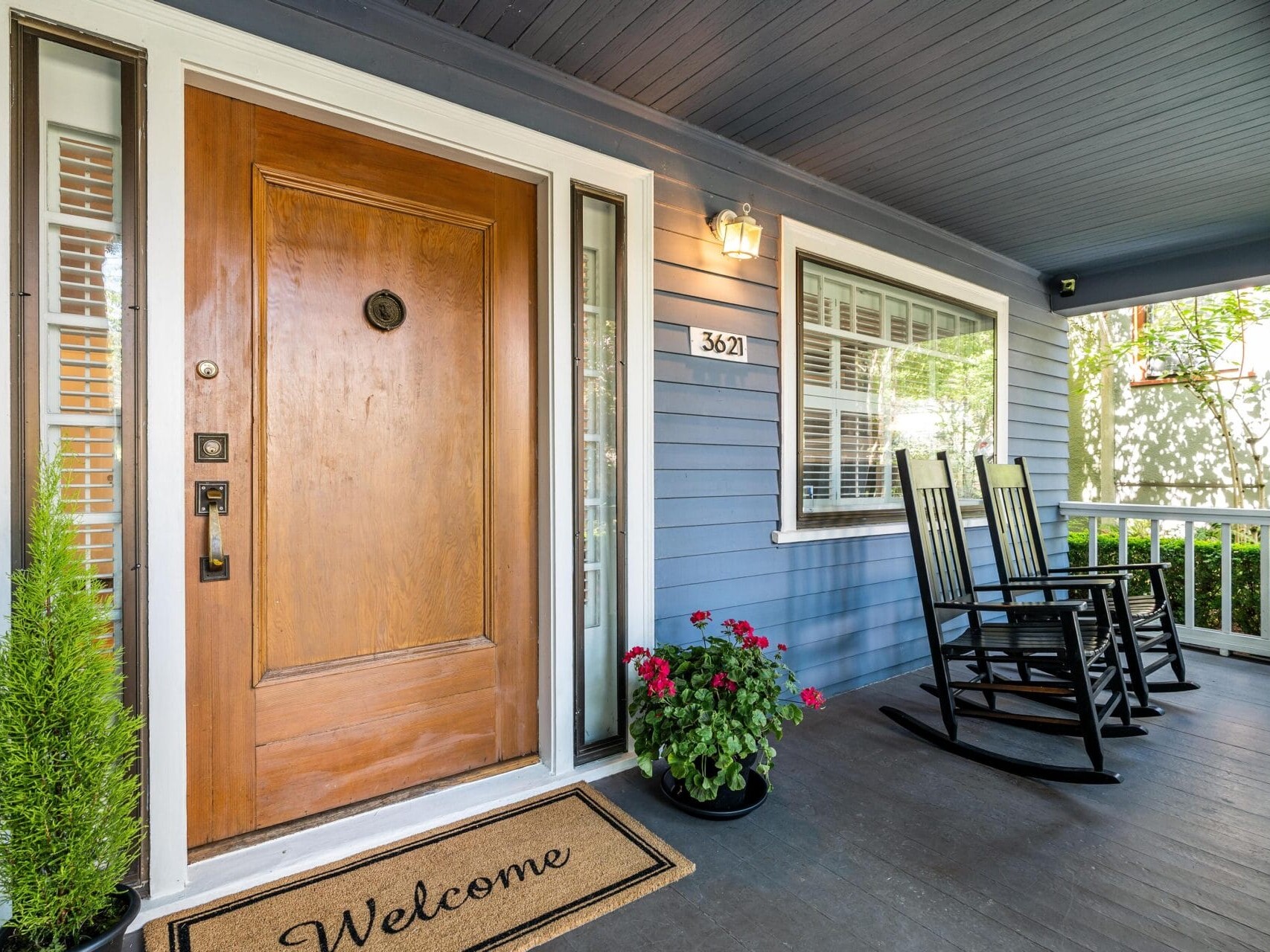 Front porch with a wooden door, flanked by sidelights and a Welcome mat. Two black rocking chairs face a potted plant with red flowers. Blue exterior walls feature a house number, 3621. A light fixture is above the door, and a white railing lines the porch.