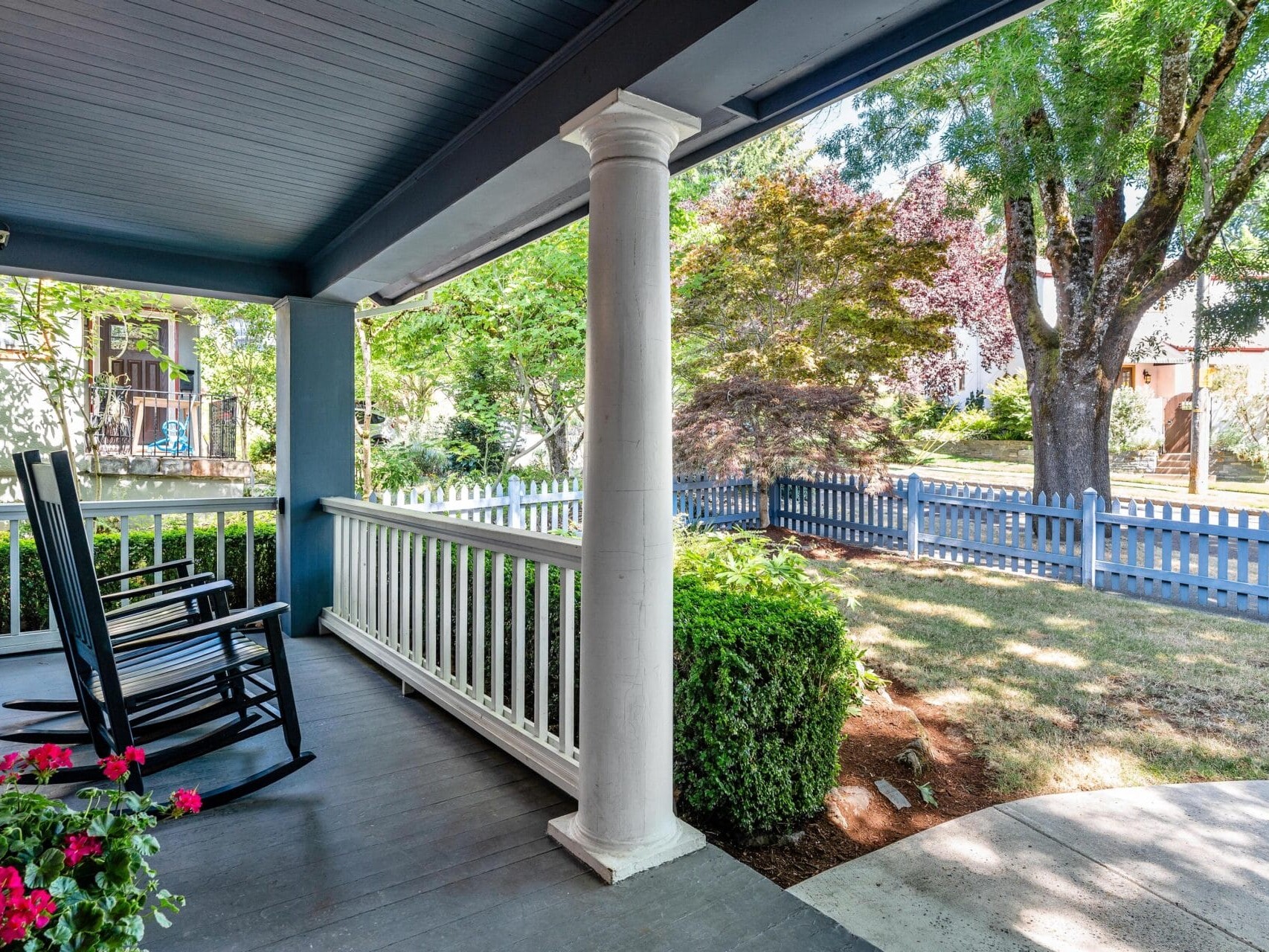A cozy porch with two black rocking chairs overlooks a lush front yard with trees and flowers. A blue picket fence borders the property, with a quiet neighborhood street beyond. Bright sunlight illuminates the peaceful scene.
