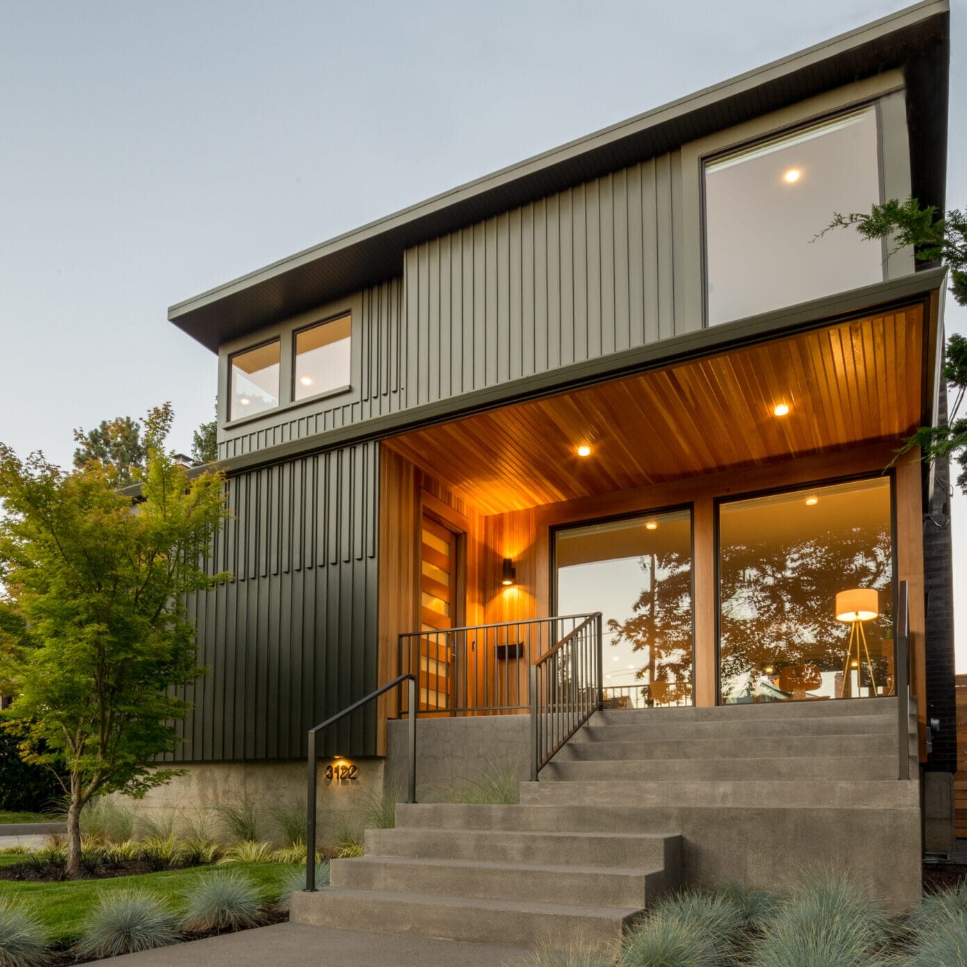 A modern two-story house in Portland, Oregon boasts large windows and a warm wooden entrance. The exterior features sleek metal siding and a concrete staircase leading to the front door of this beautifully landscaped property, surrounded by neatly arranged greenery and trees.