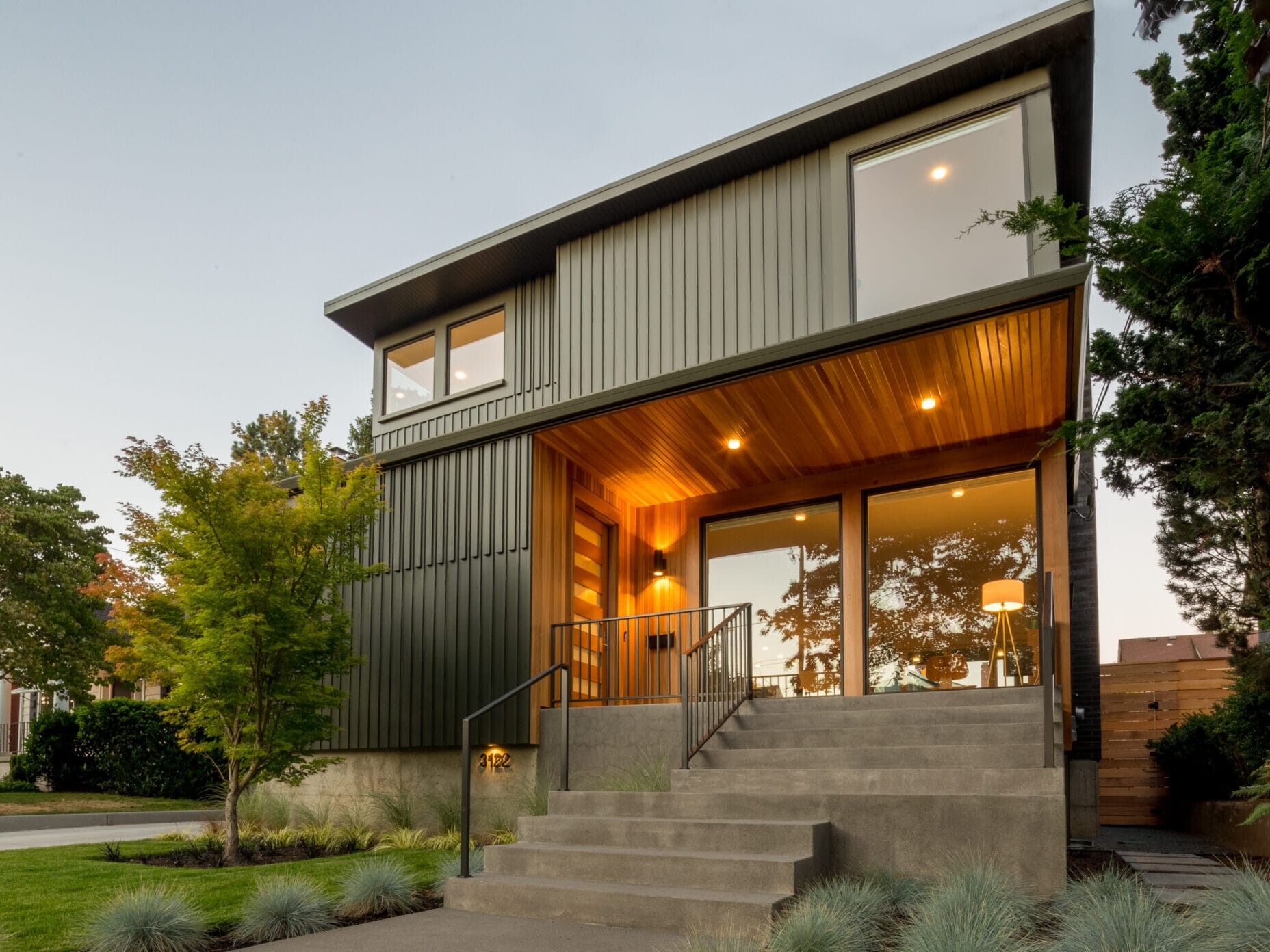 A modern two-story house in Portland, Oregon boasts large windows and a warm wooden entrance. The exterior features sleek metal siding and a concrete staircase leading to the front door of this beautifully landscaped property, surrounded by neatly arranged greenery and trees.