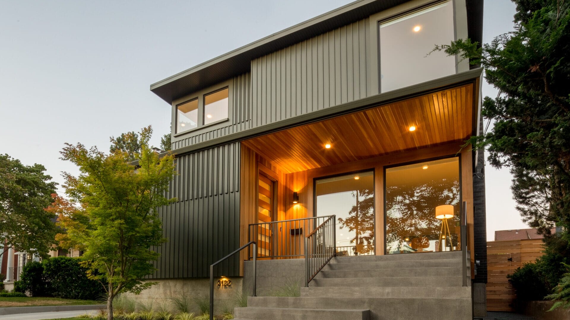 A modern two-story house in Portland, Oregon boasts large windows and a warm wooden entrance. The exterior features sleek metal siding and a concrete staircase leading to the front door of this beautifully landscaped property, surrounded by neatly arranged greenery and trees.