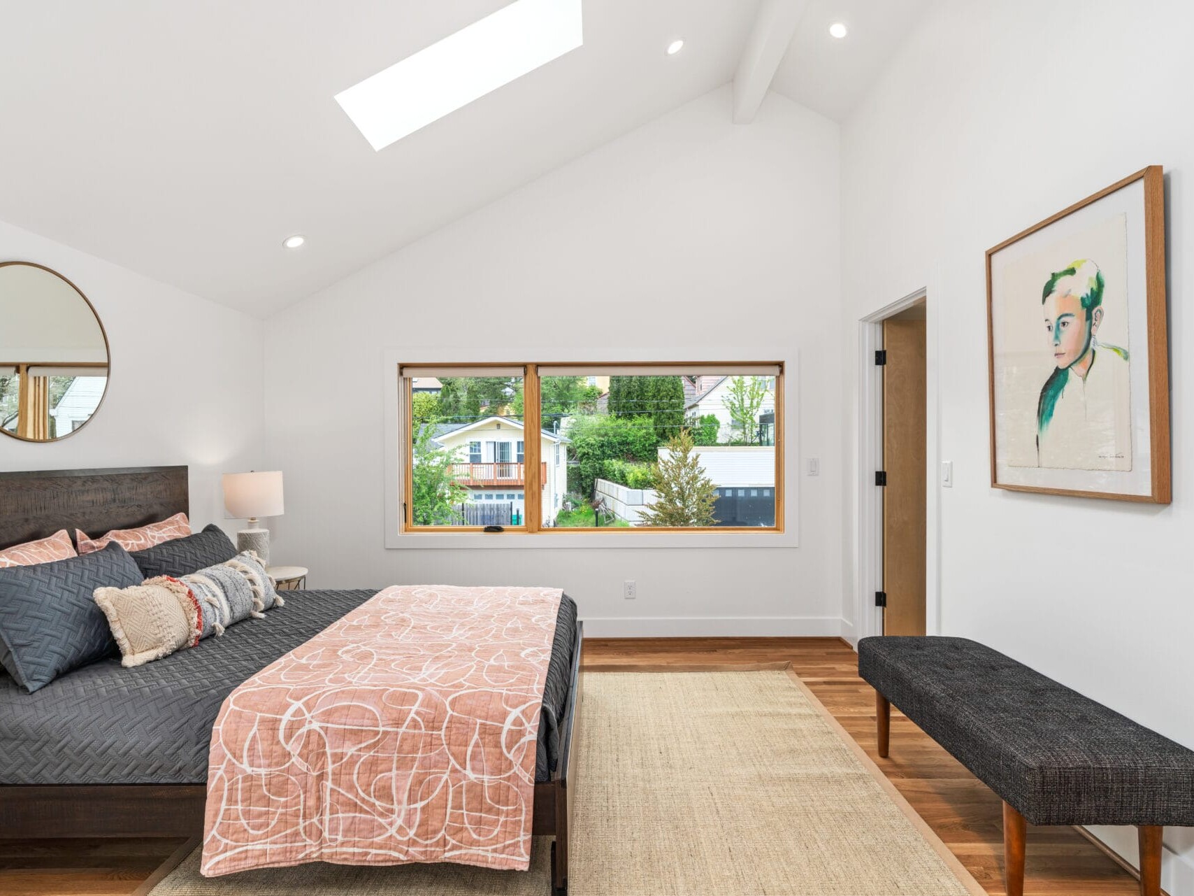 A modern Portland bedroom with a sloped ceiling showcases a dark wooden bed adorned with patterned pillows and blankets. A large window offers an idyllic Oregon view, while a round mirror, lamp on a bedside table, bench, and framed artwork complete the serene space.