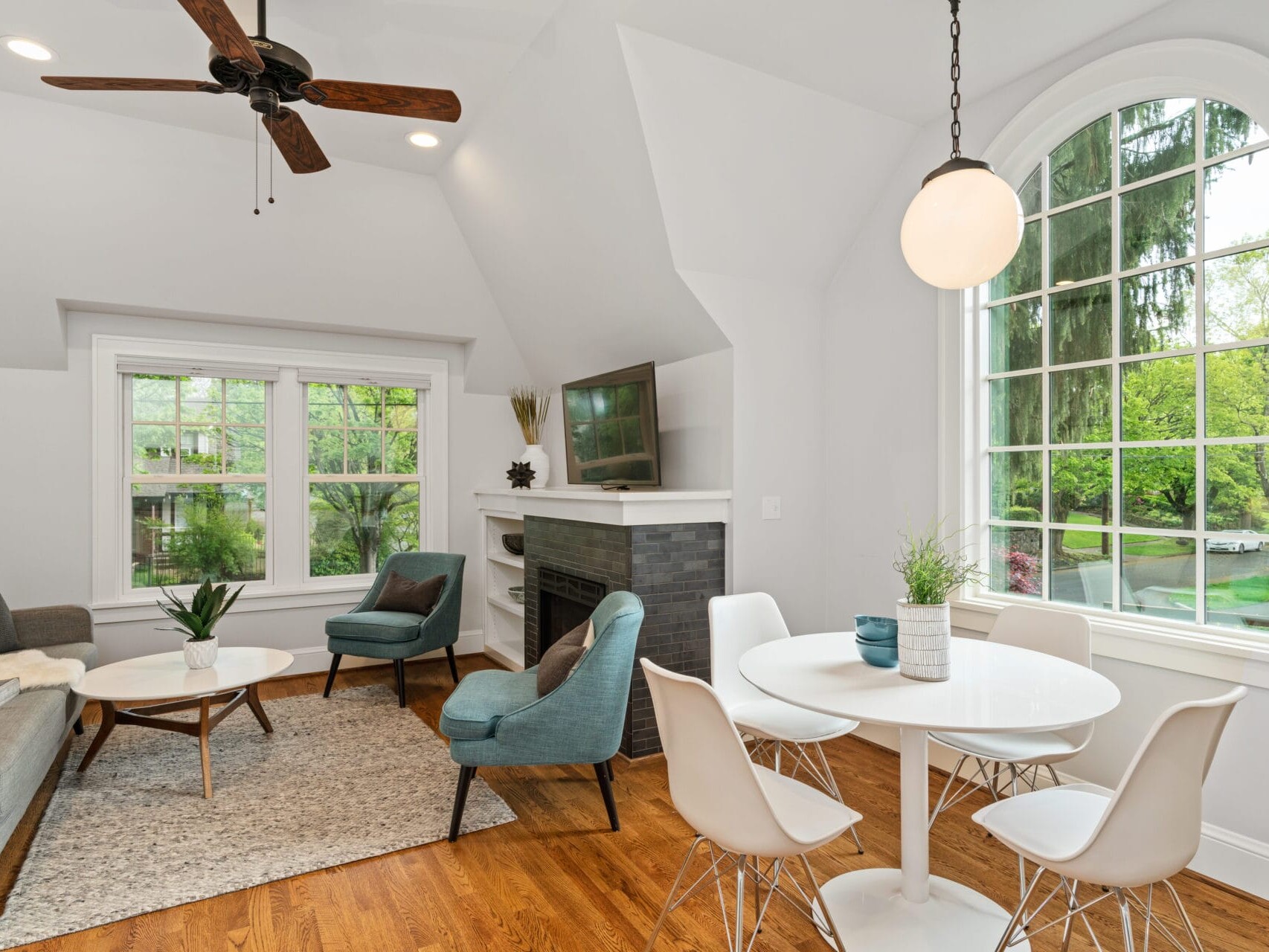 A bright living room in Portland, Oregon, showcases a modern design with a gray sofa, blue chairs, and a round white dining table. A ceiling fan and arched window enhance the space, while a flat-screen TV sits above a fireplace adorned with black tiles.