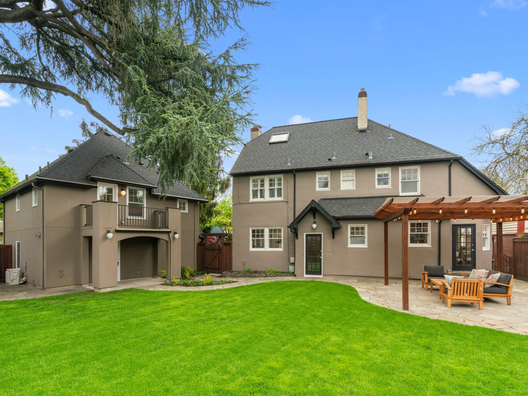 Rear view of a large gray two-story house in Portland, Oregon, featuring a balcony, adjacent small structure, and spacious patio. A pergola with seating graces the well-maintained lawn in the backyard, while tall trees frame the scene under a clear blue sky.