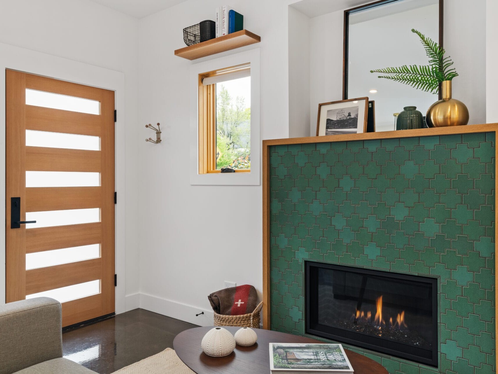 A cozy living room in Portland, Oregon, features a modern green tiled fireplace. A wooden door with narrow horizontal windows is on the left. The mantel displays decor including a plant and framed photos. A small window above a chair lets in natural light, while a table holds books.