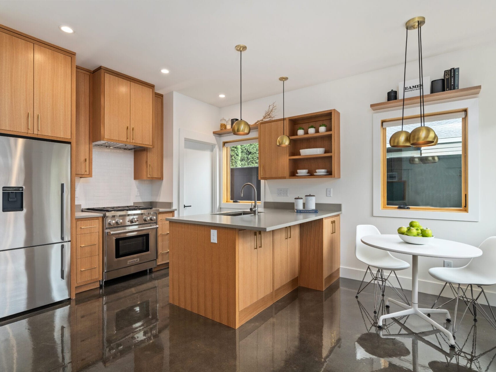 A modern kitchen with wood cabinets, stainless steel appliances, and a central island with a sink. Two pendant lights hang above the island. A small dining table with white chairs and a bowl of green apples is near the window.