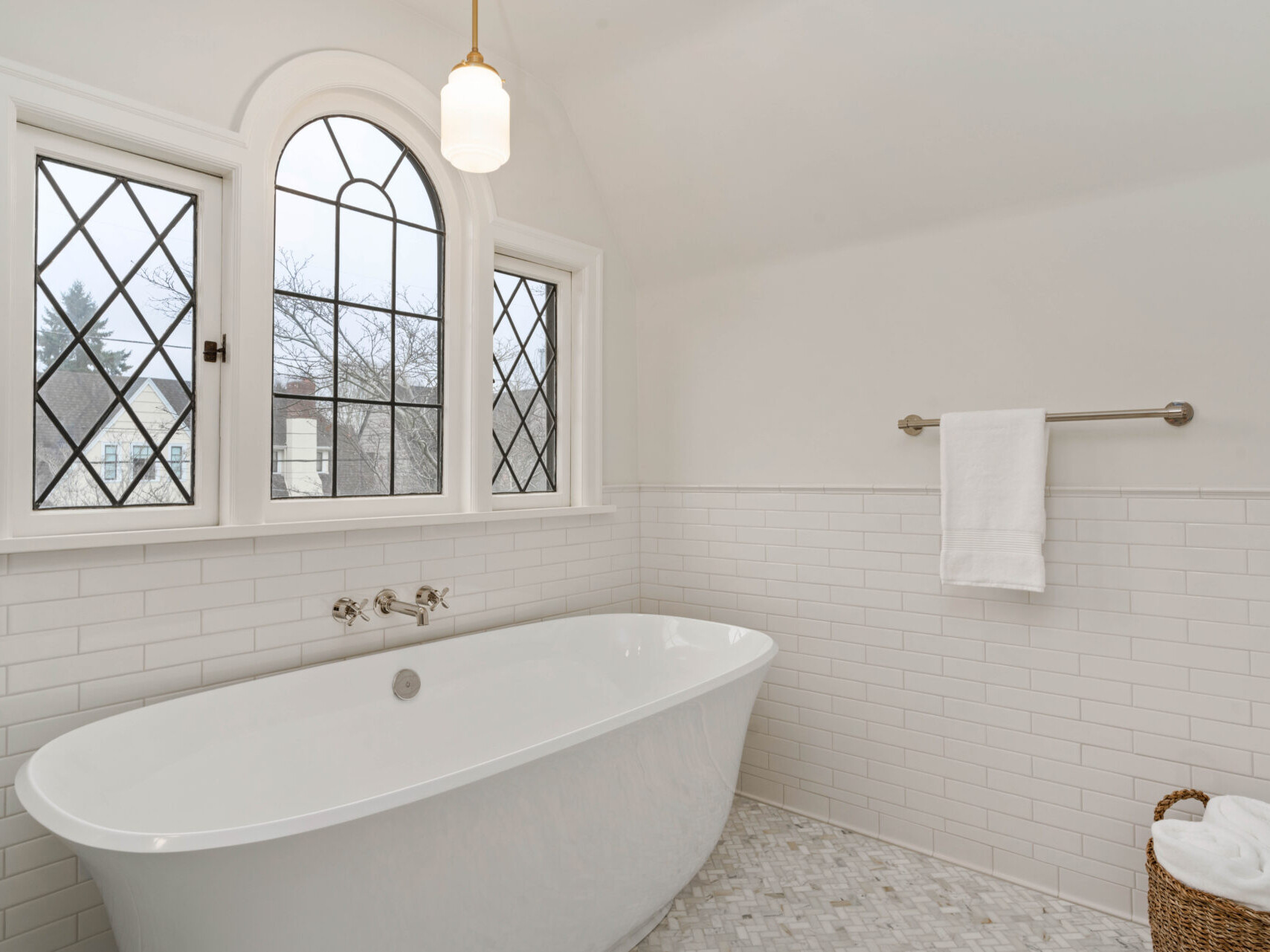 A bright bathroom in Portland, Oregon features white subway tile walls and a freestanding bathtub below two arched windows with diamond-patterned panes. A white towel hangs on a rail, and a basket with towels rests on the mosaic tile floor. A pendant light hangs overhead.