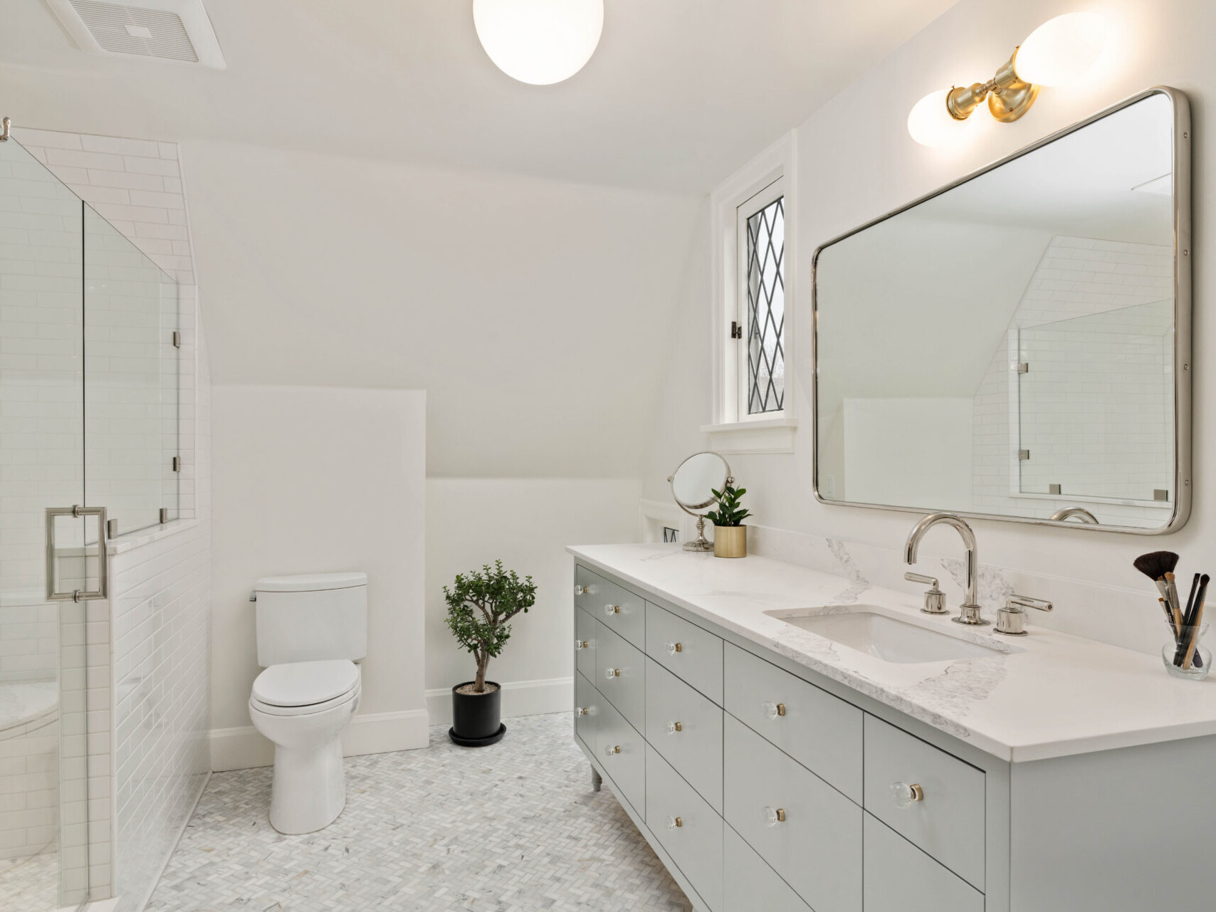 A modern, white bathroom in Portland, Oregon features a large mirror above a double sink with a marble countertop. A small potted plant sits between the toilet and vanity. A glass-enclosed shower is on the left, and the room is well-lit with natural and ceiling light.
