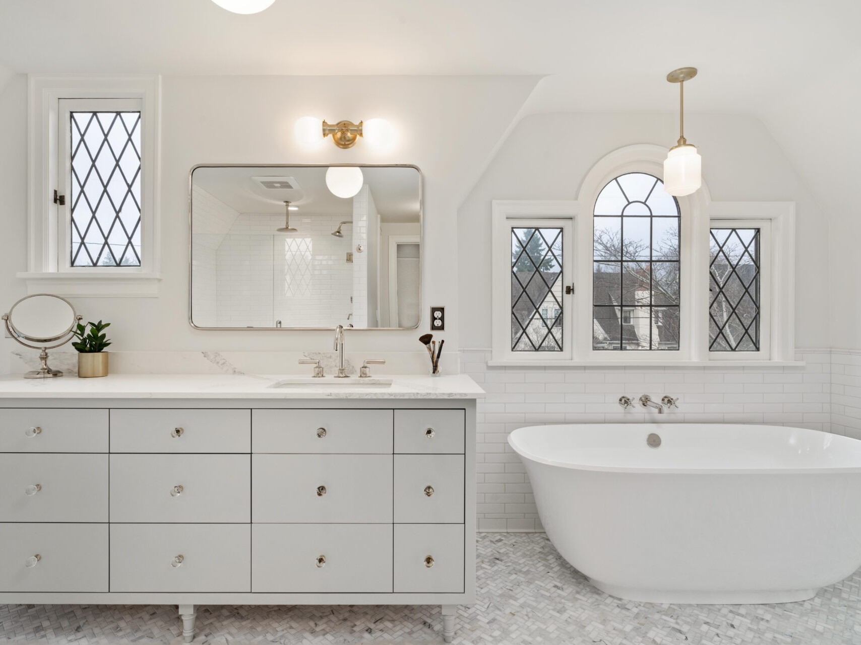 A bright, elegant bathroom in Portland, Oregon, showcases white walls and a freestanding bathtub beneath arched windows. On the left, a wide mirror and vanity with drawers feature a small round mirror and a potted plant. The floor displays a light mosaic pattern.
