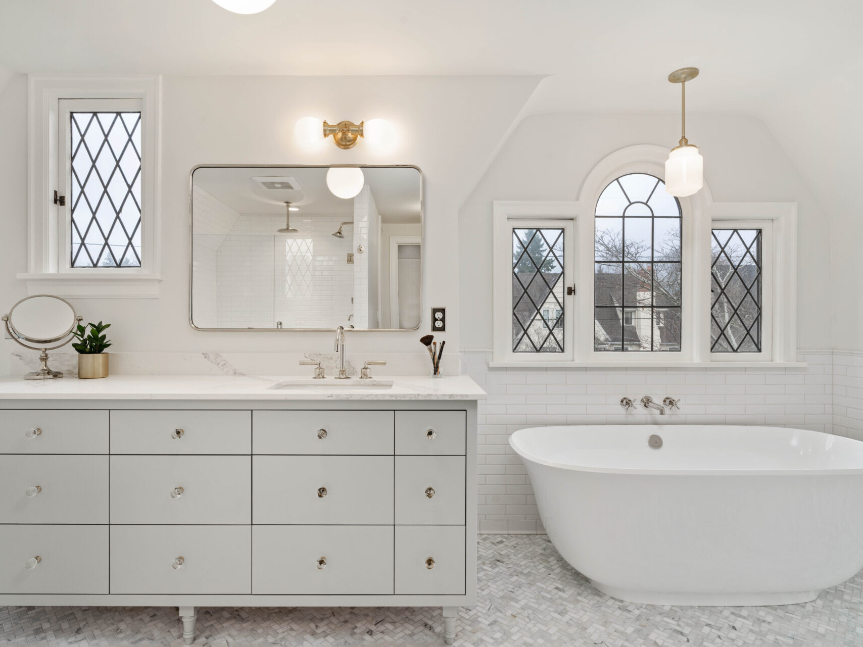 A bright, elegant bathroom in Portland, Oregon features a freestanding white bathtub and a large mirror above a light gray vanity with drawers. Decorative windows, white walls, and a potted plant complement the space. A small round mirror on the vanity and light patterned tile complete the look.